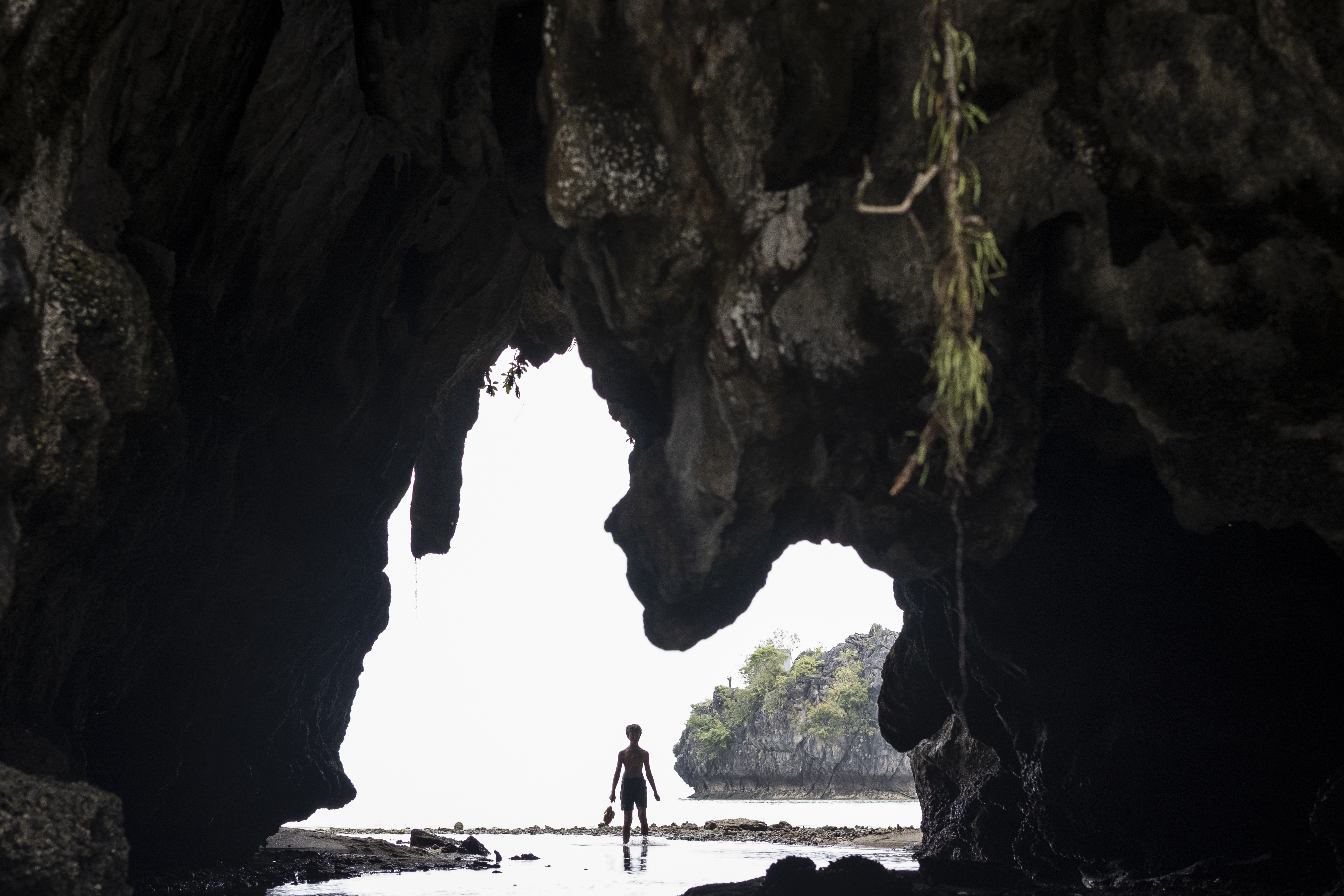 "Silhouette of a person standing at the entrance of a large cave, looking out toward a bright, rocky shoreline with water and distant vegetation, creating a dramatic contrast between the dark cave and bright exterior."