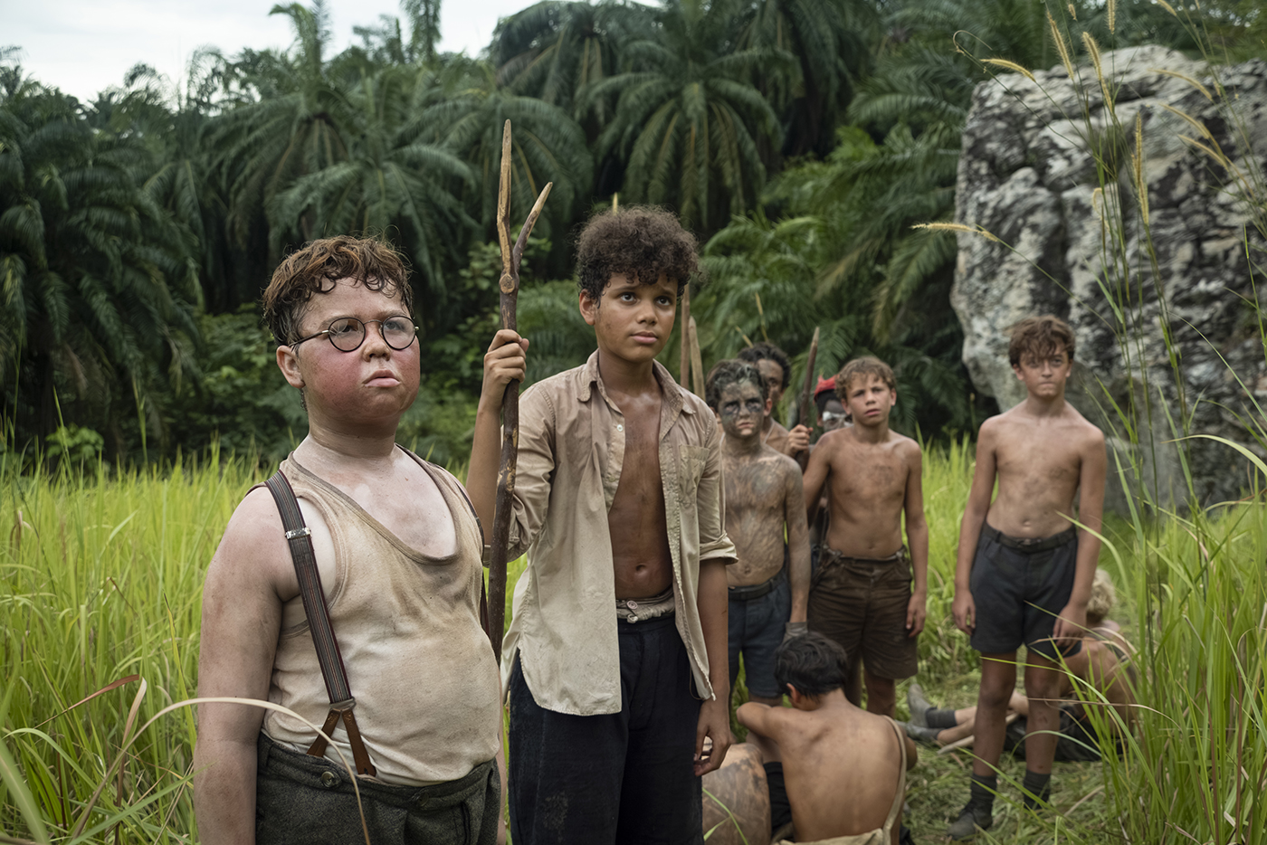 "A group of dirty, shirtless boys stand in tall grass with jungle foliage and a rock behind them. Some hold makeshift spears, appearing serious and alert, as if stranded or surviving in the wild."