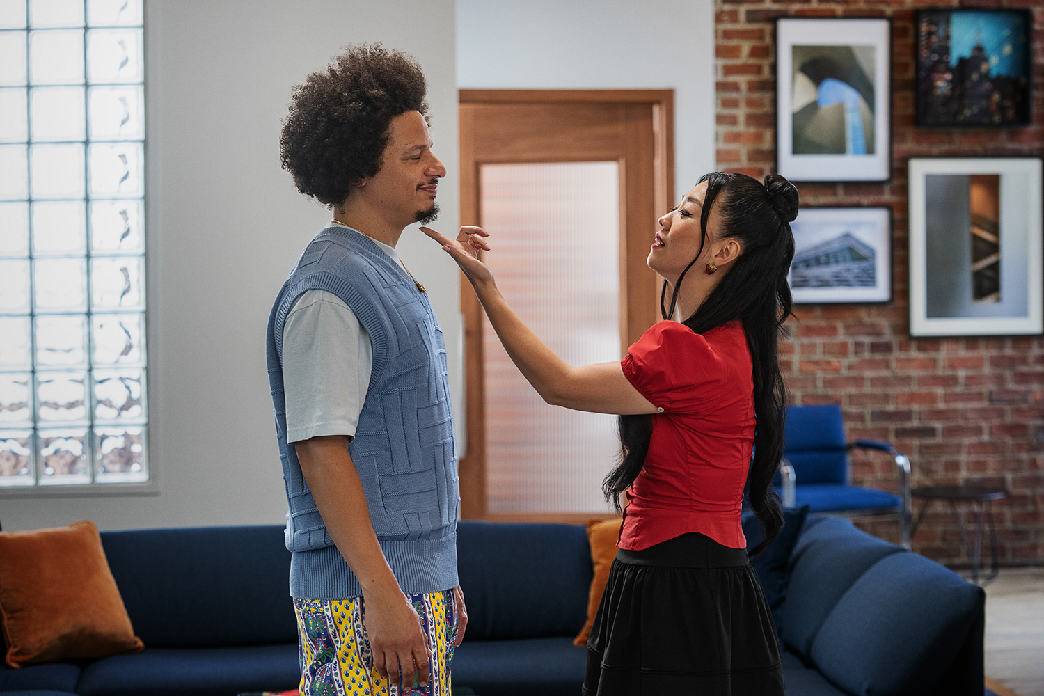 "A man and woman stand facing each other in a modern living room with brick walls, framed pictures, a blue sofa and orange pillows. The woman is touching the man's chin, both are seen having an emotional conversation."
