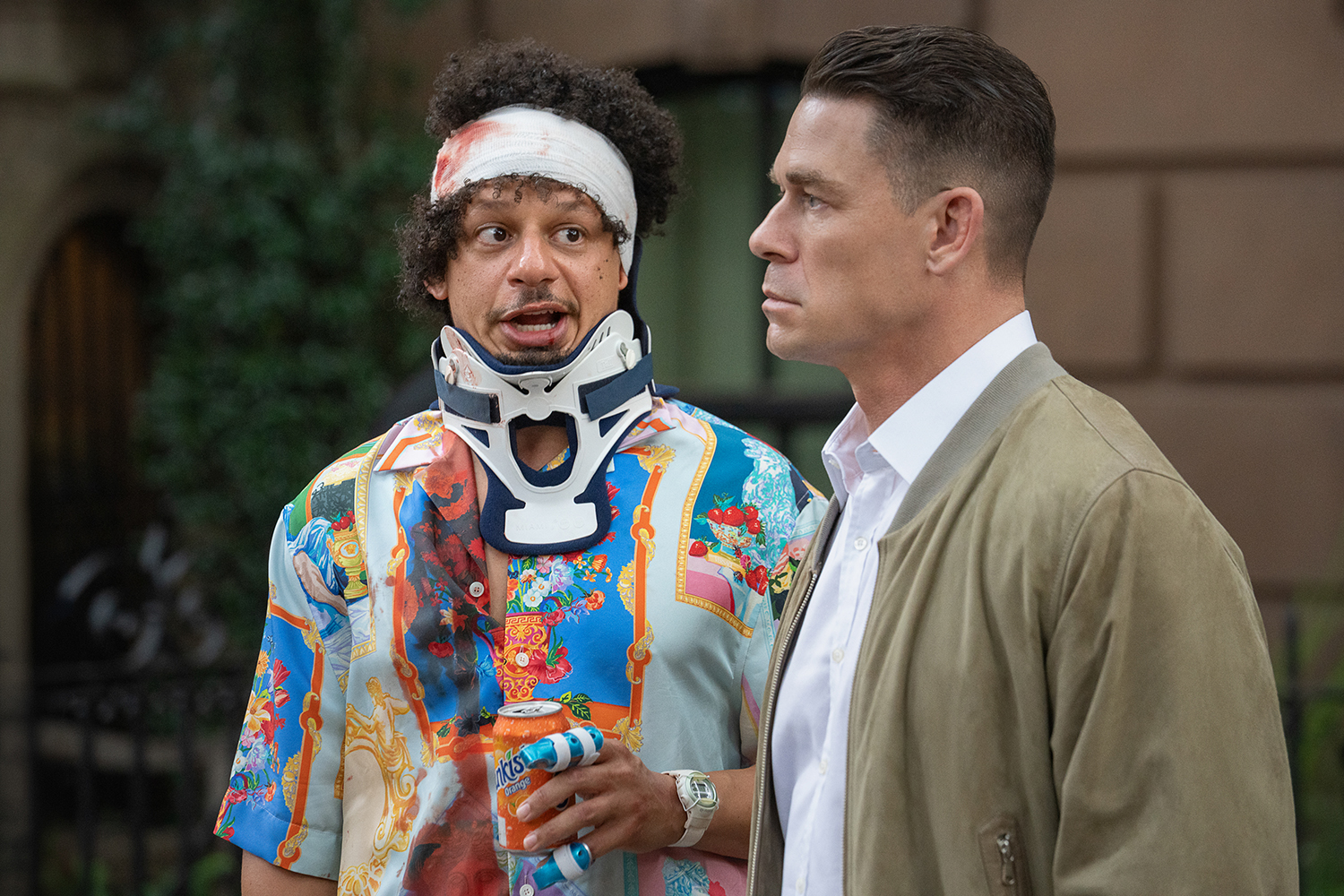 "Two men outdoors: one wears a colorful shirt, head bandage, and neck brace, holding a can, looking surprised; the other in a tan jacket and white shirt, looking serious. Urban background with greenery and brownstone building."