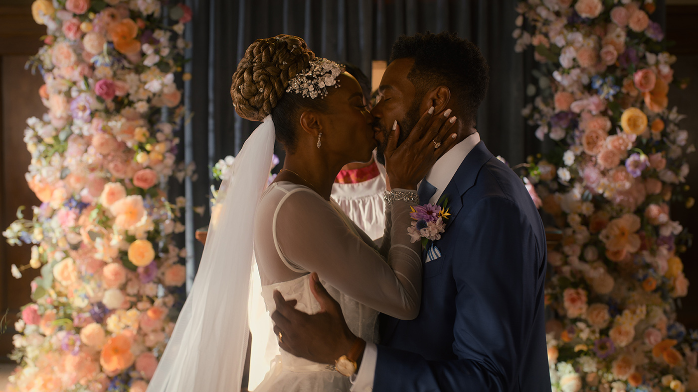"The bride and groom share a kiss at their wedding ceremony surrounded by colorful flower arrangements and soft lights, capturing a joyful and romantic moment."