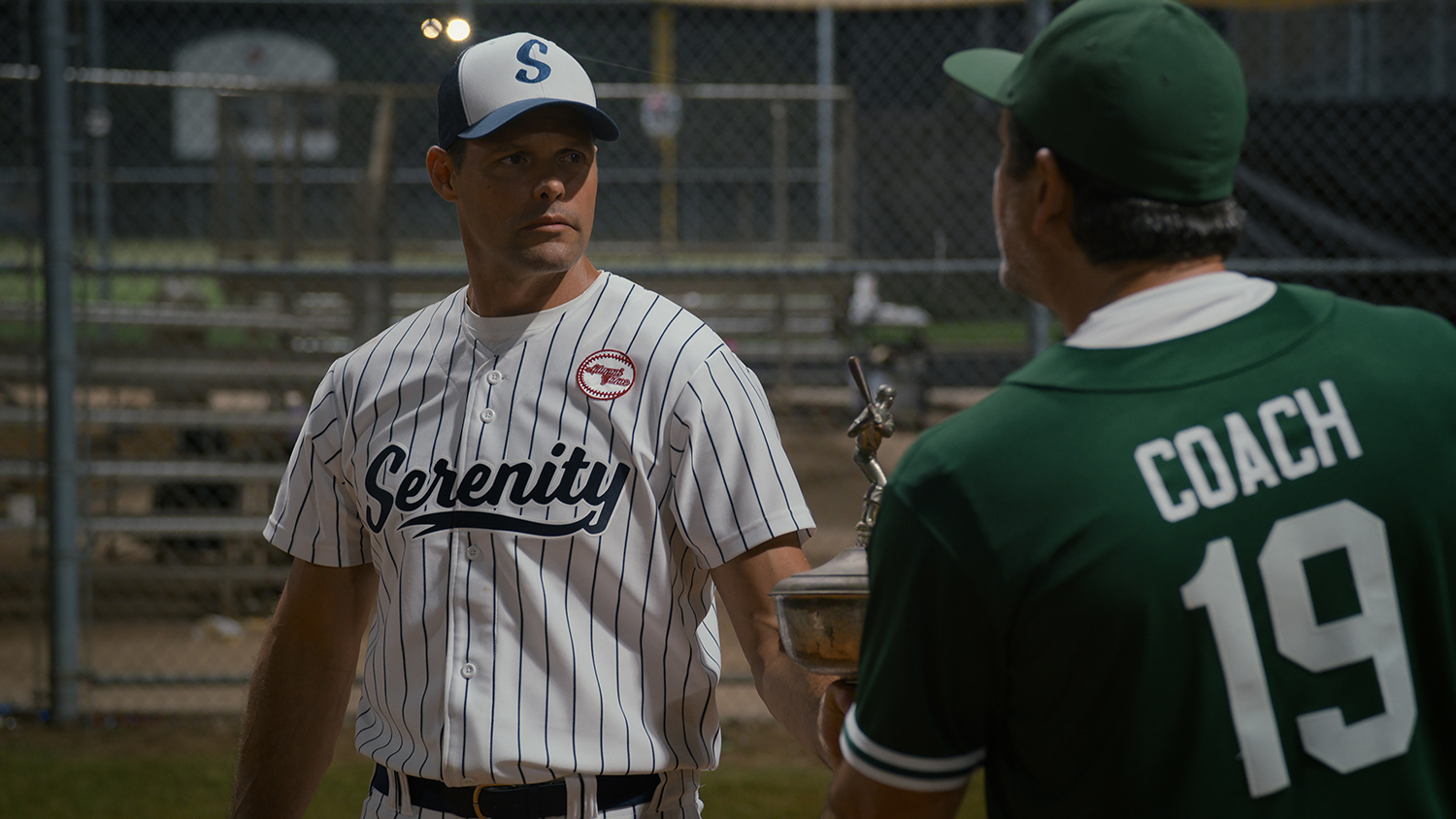 "Two men on a baseball field at night, one in white pinstripes "Calmness" Uniform and cap, second green jersey labeled "coach 19," Holding a trophy, with empty bleachers and a chain link fence in the background."