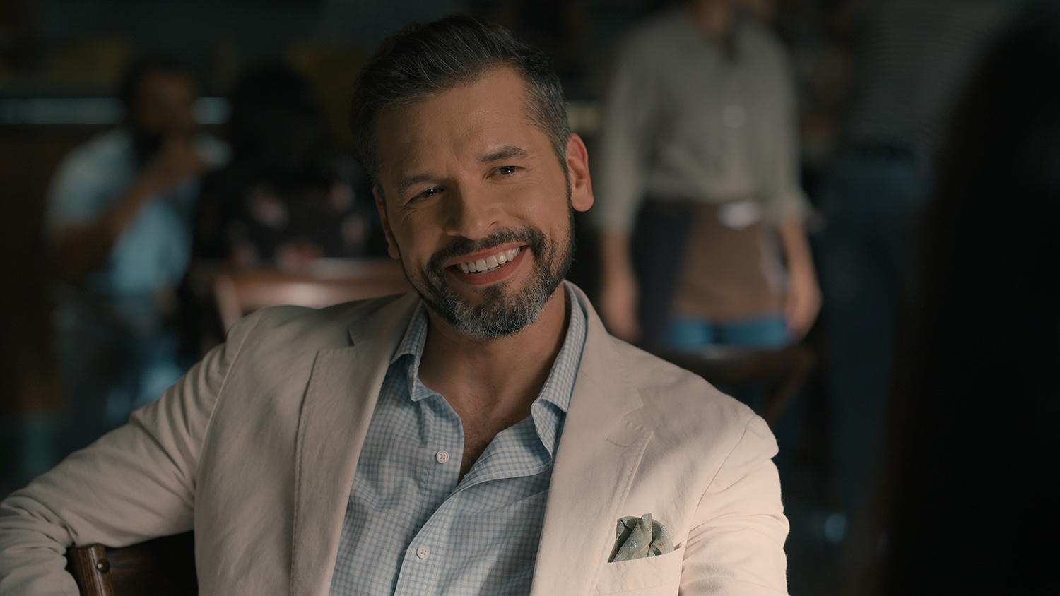 "A bearded man, wearing a light blazer and checked shirt, is smiling while sitting at a table in a dimly lit café, with blurry people and a casual atmosphere in the background."