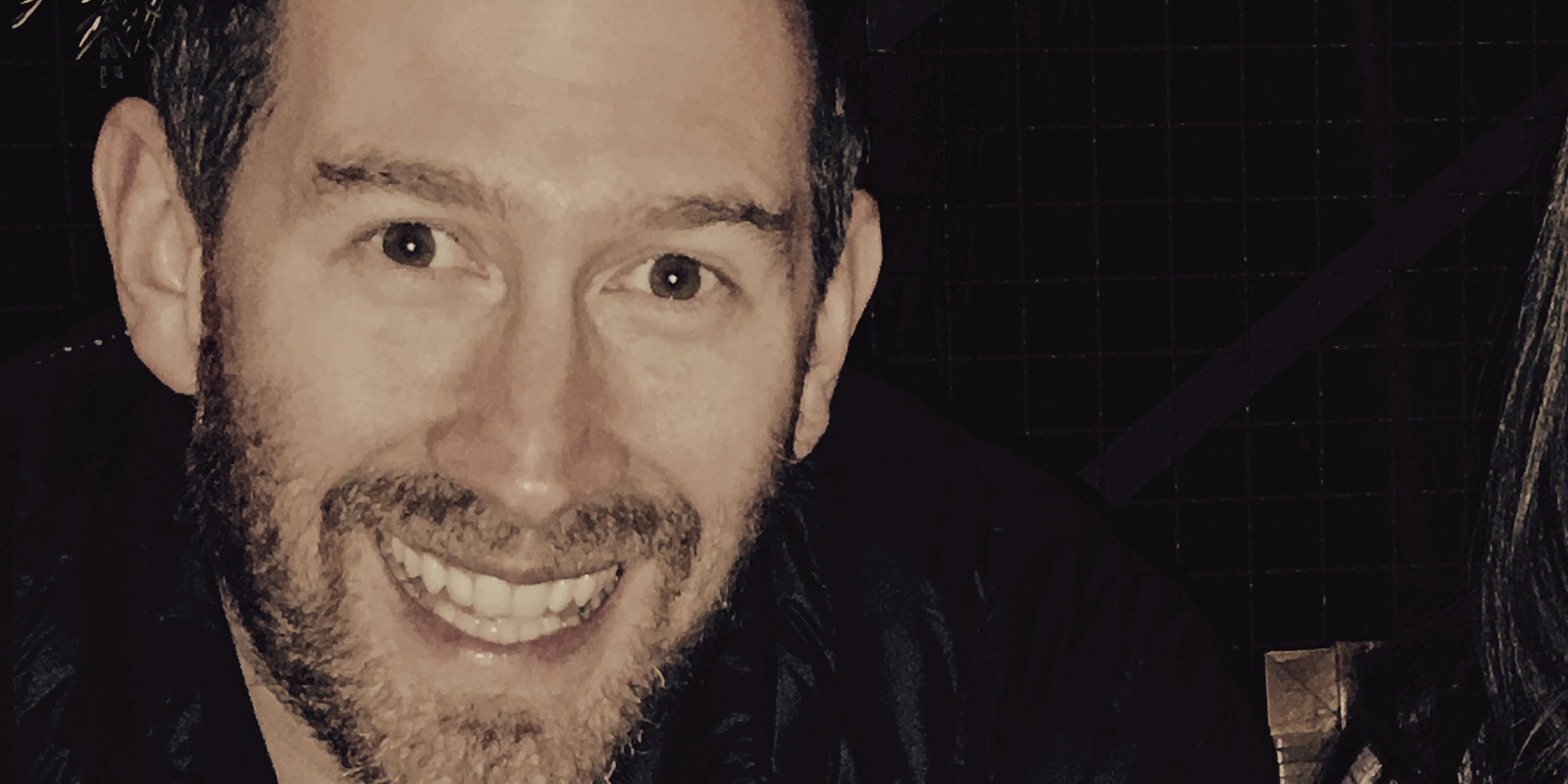 "Smiling man with short hair and beard in a dark indoor setting, with a grid-patterned wall in the background."