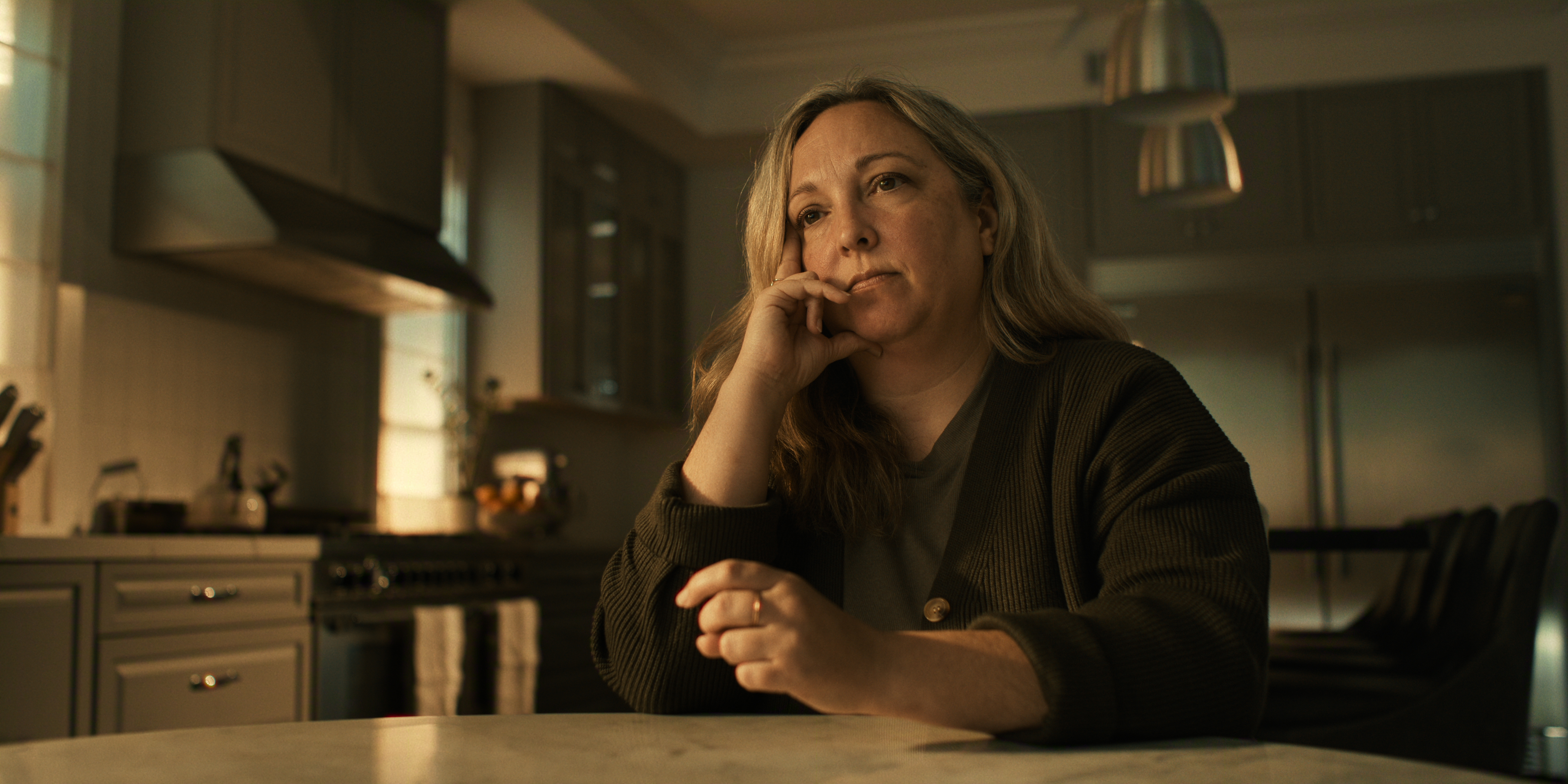 "Woman sitting at kitchen table, resting her chin on her hand, looking thoughtful. The kitchen is modern with gray cabinets, stainless steel appliances, and warm indoor lighting."