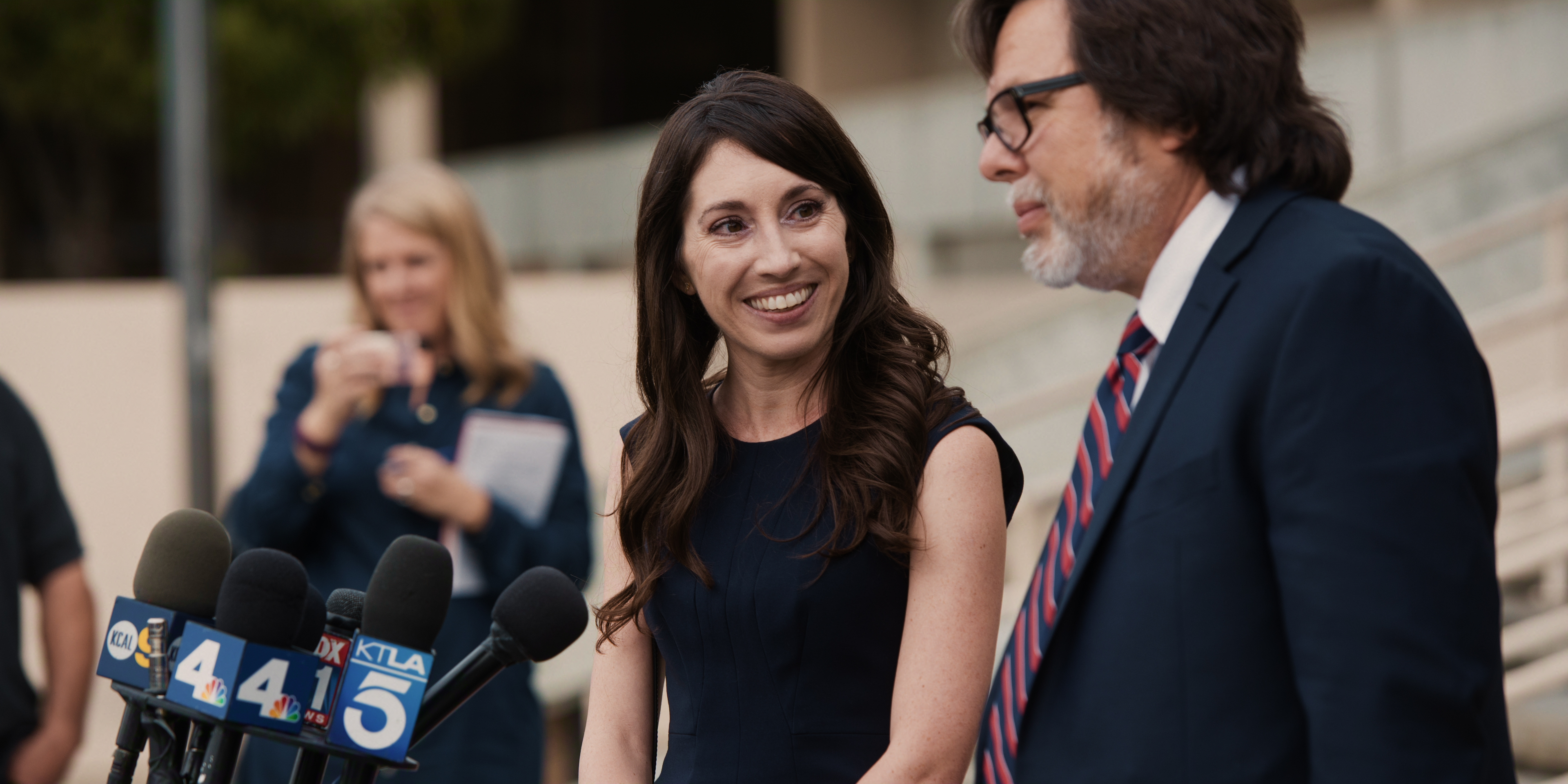 "A woman and a man stand in front of a microphone with a news station logo, smiling and talking on a staircase outside. Reporters and news crews are visible in the background, creating a professional and public setting."