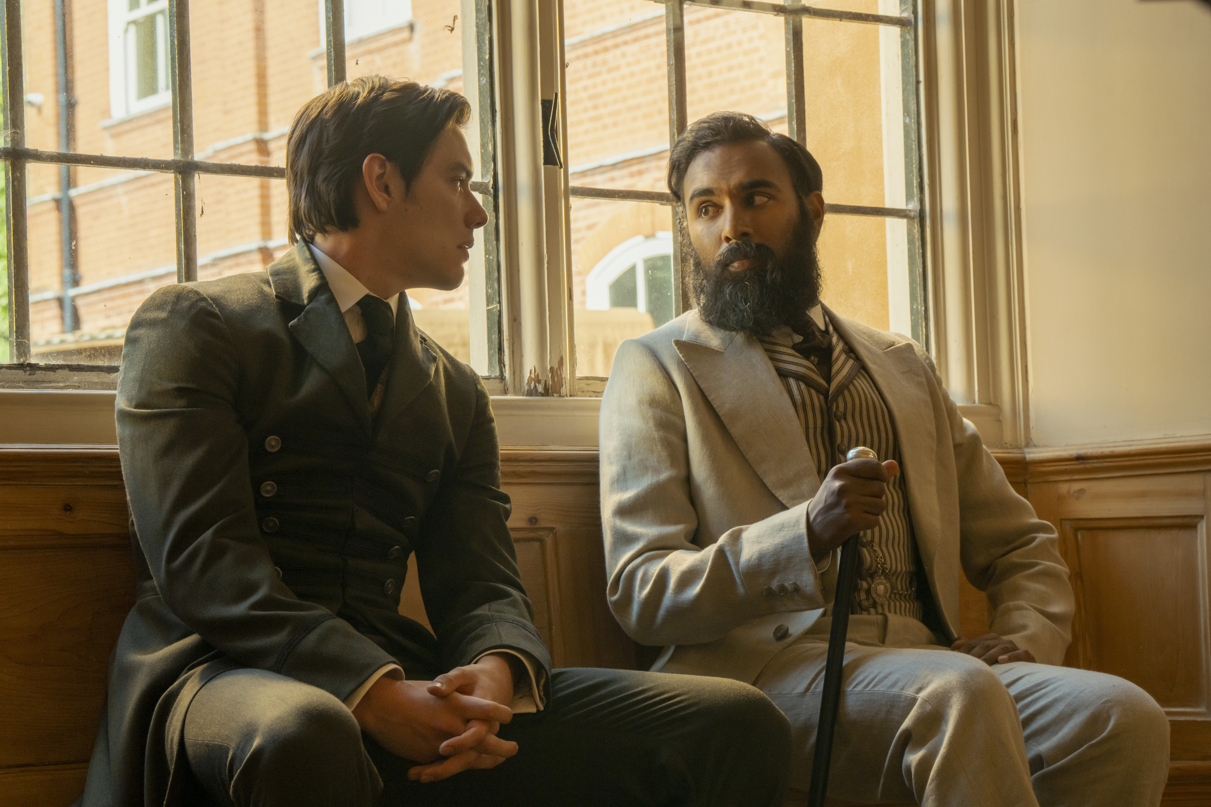 &Quot;Two Men In Period Clothing Sit On A Wooden Bench By A Large Window In A Sunlit Room, Engaged In Conversation. One Wears A Dark Suit, The Other A Light Suit And Holds A Cane, With Brick Buildings Visible Outside The Window.&Quot;