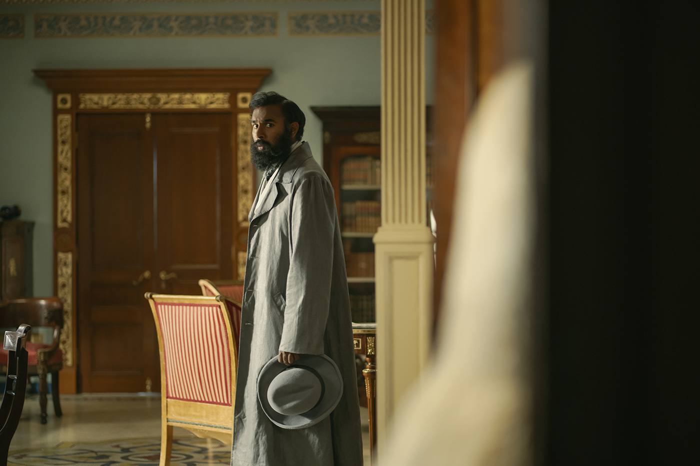 &Quot;Man In A Long Coat Holding A Hat Stands In An Elegant, Ornate Room With Wooden Door, Classical Columns, Striped Chairs, And Bookshelves, Looking Contemplative. Warm Daylight Fills The Sophisticated, Historical Setting.&Quot;