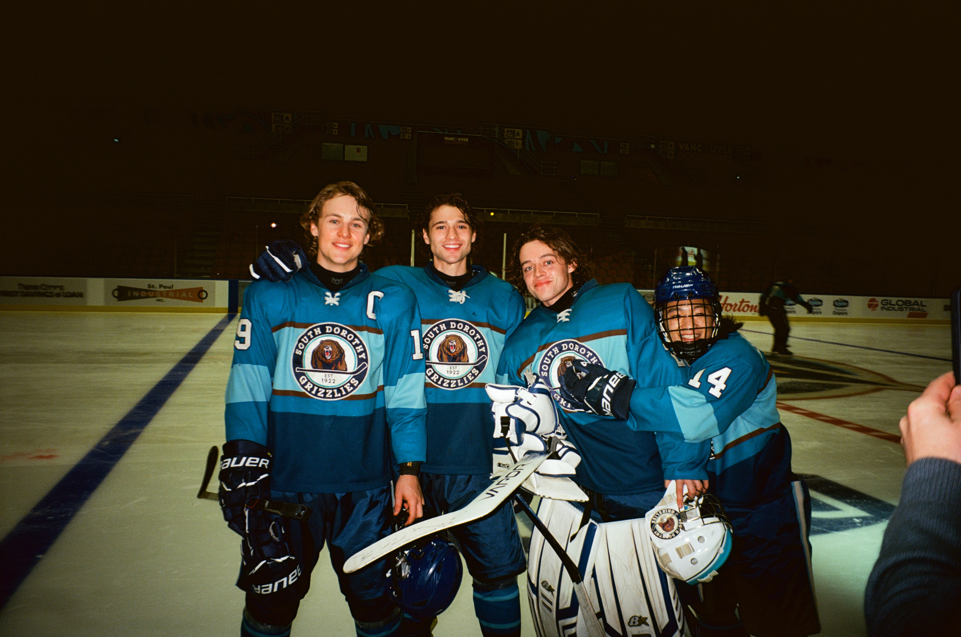 &Quot;Four Hockey Players In Blue Uniforms Posing Together On An Ice Rink, Smiling For A Photo. The Rink Is Indoors With Boards And Advertisements Visible In The Background, Suggesting A Team Game Or Practice Session.&Quot;