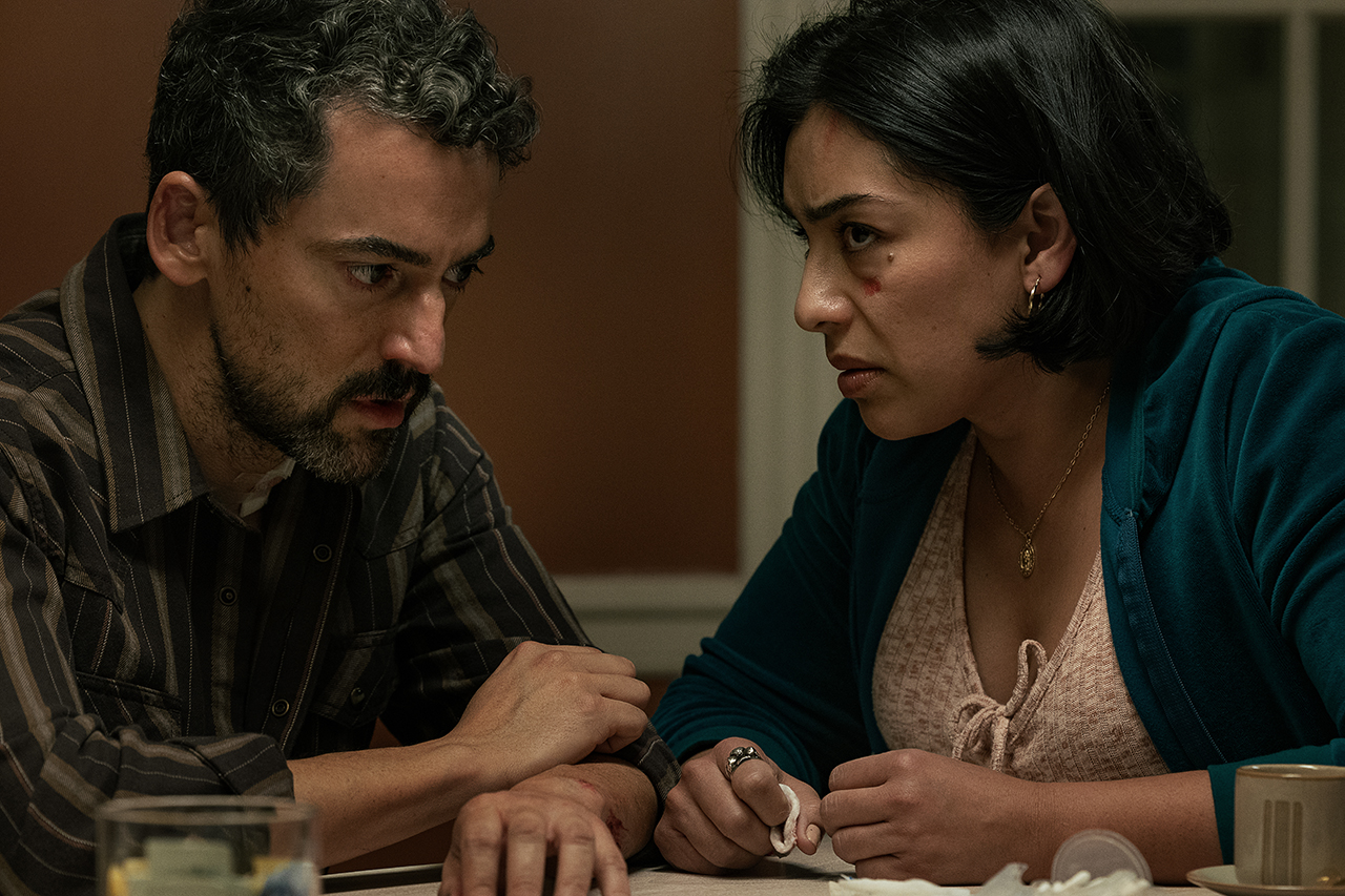 &Quot;Two Concerned Adults Sit Closely At A Table In A Dimly Lit Room, Engaged In A Serious Conversation. The Setting Is A Domestic Environment With A Warm-Toned Wall And Household Items Present, Suggesting An Intense Or Emotional Moment.&Quot;