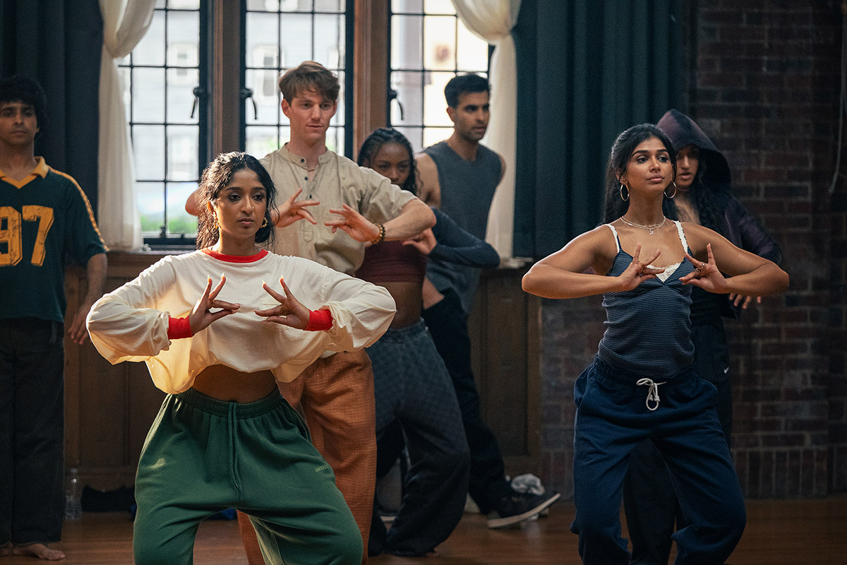 "A diverse group of young adults practicing dance in a studio with wooden floors, brick walls, and large windows. The two women in the front are focused on their hand movements, suggesting a choreographed routine."