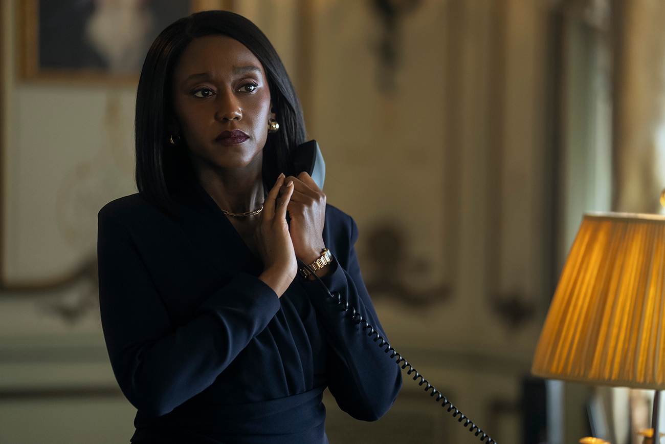 &Quot;Woman In Formal Attire Stands In An Elegant, Softly Lit Room, Holding A Corded Phone To Her Ear, Appearing Thoughtful Or Concerned. A Table Lamp And Ornate Decor Are Visible In The Background.&Quot;