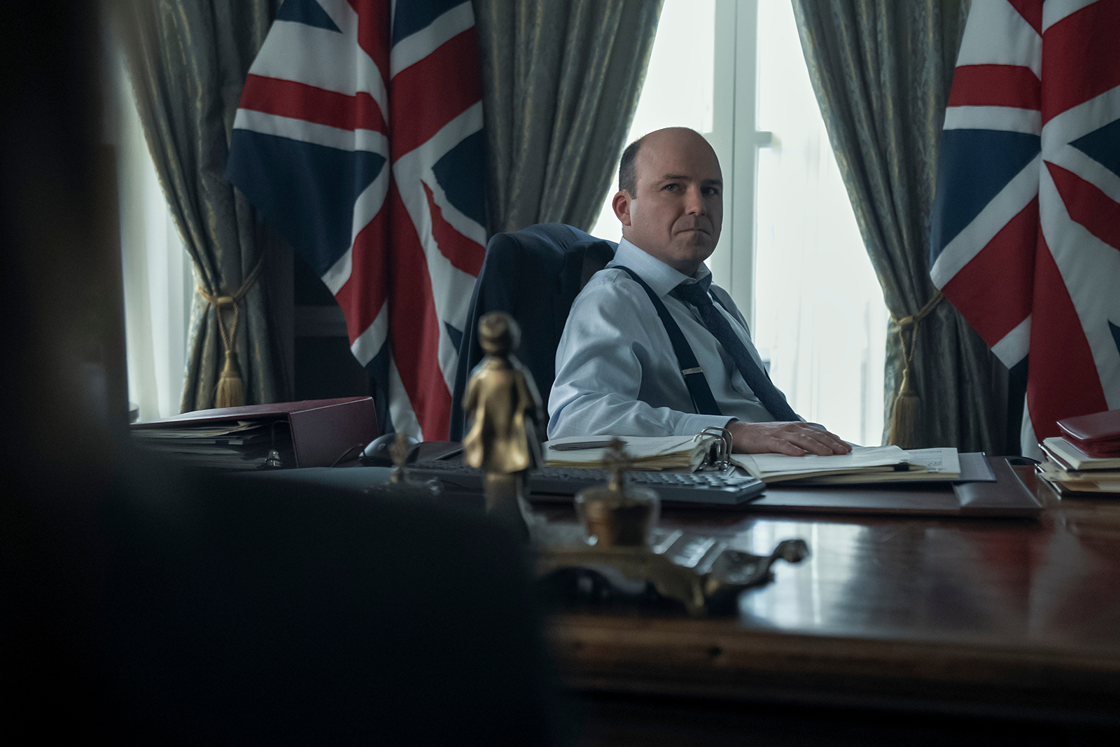&Quot;A Man In A Formal Office Sits At A Large Desk With Papers, Backed By British Flags And Heavy Curtains, Appearing To Be In A Position Of Authority Or Government.&Quot;