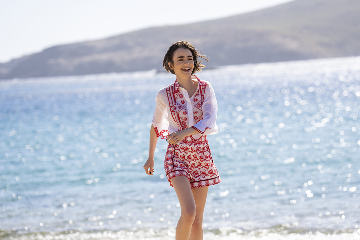 &Quot;A Young Woman In A Patterned Shirt And Shorts Smiles While Walking Along A Sunny Beach, With Sparkling Blue Water And Distant Hills In The Background.&Quot;