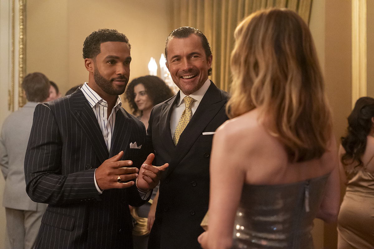 &Quot;Two Men In Suits And A Woman In A Metallic Dress Converse At An Elegant Indoor Party, Surrounded By Other Formally Dressed Guests In A Well-Lit, Upscale Setting With Gold Accents And Curtains.&Quot;