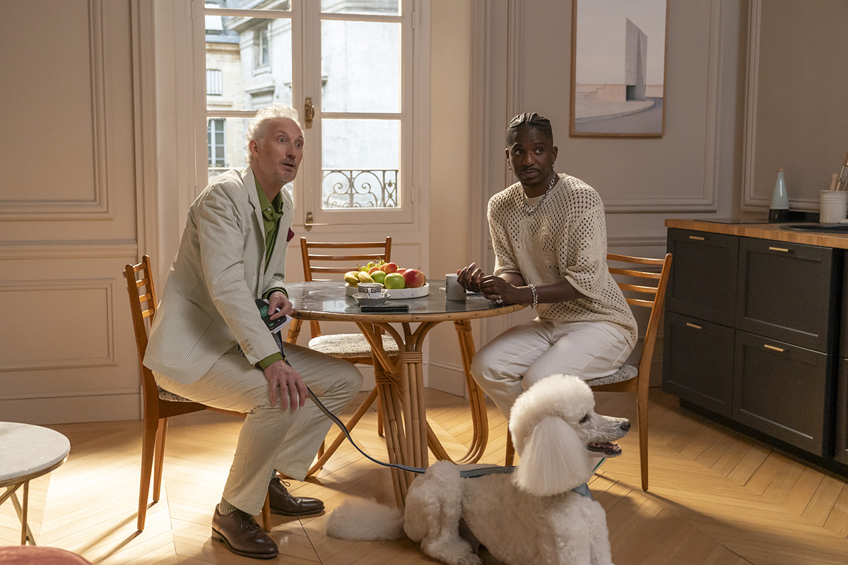 &Quot;Two Men Sitting At A Round Wooden Table In A Bright, Elegant Apartment With A Window, Fruit Bowl, And Modern Decor. A White Poodle On A Leash Sits On The Floor Beside Them.&Quot;