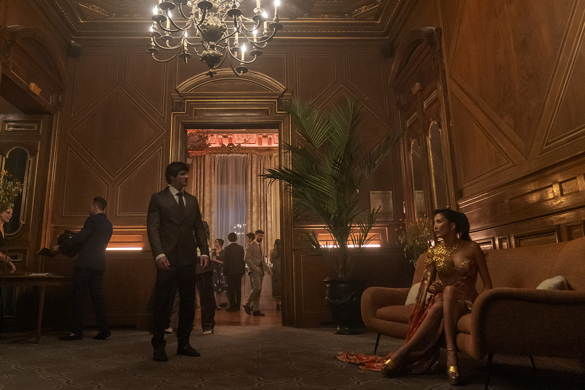 &Quot;A Man In A Suit Stands In An Elegant, Wood-Paneled Lounge With A Chandelier, While A Woman In A Gold Dress Sits On A Couch; Other People Are Visible In The Background Socializing At A Formal Event.&Quot;