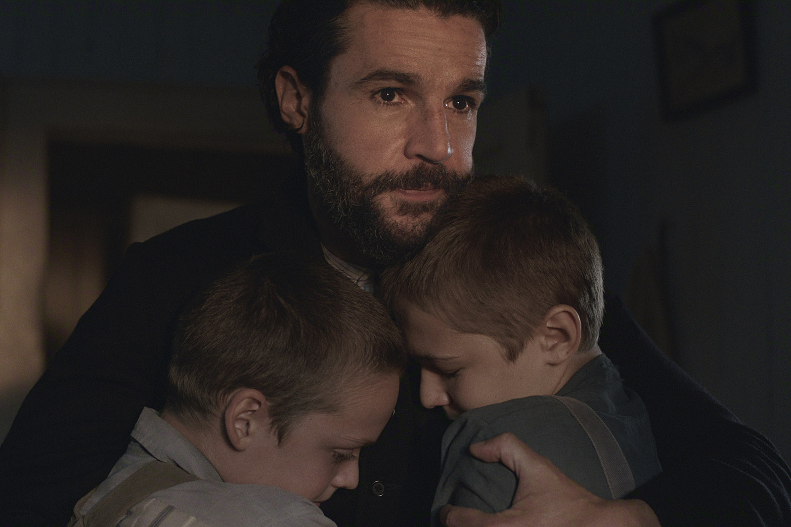&Quot;A Bearded Man In A Dimly Lit Room Hugs Two Young Boys Close To His Chest, Providing Comfort. All Three Have Serious, Emotional Expressions, Suggesting A Moment Of Sadness Or Reassurance In A Home Setting.&Quot;