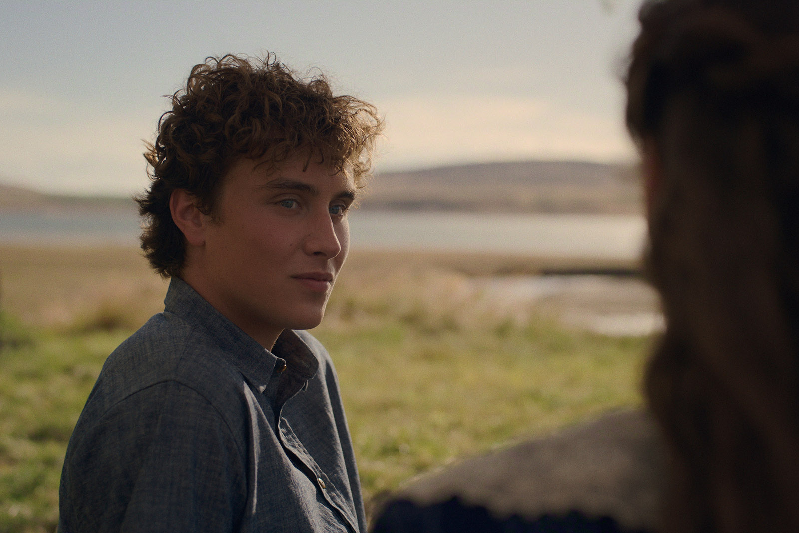 &Quot;Young Man With Curly Hair Outdoors In A Grassy Field Near Water, Wearing A Blue Shirt, Looking At A Person With Long Hair Whose Back Is To The Camera, With Hills And A Calm Sky In The Background.&Quot;