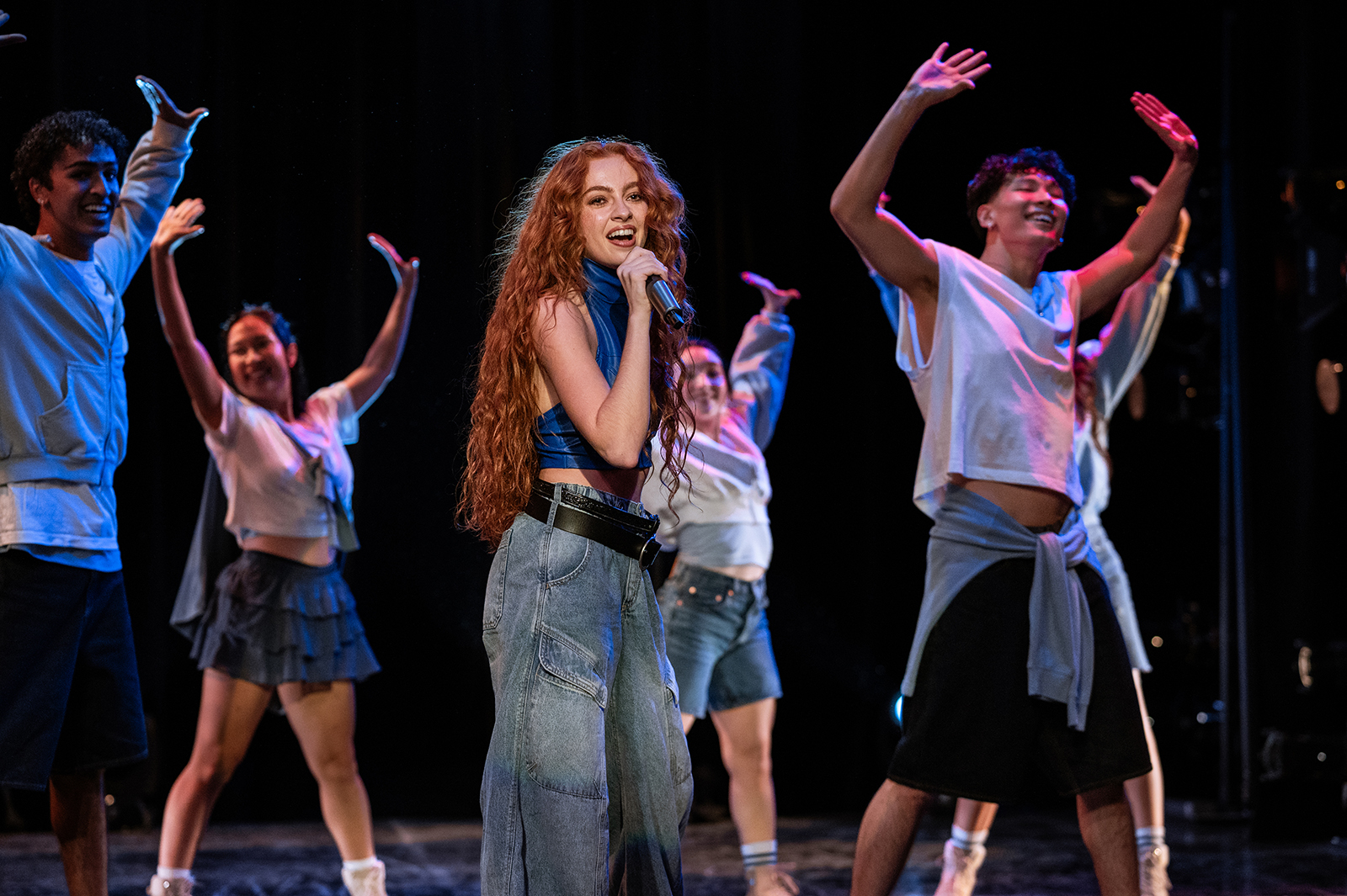&Quot;A Young Woman With Long Red Hair Sings Into A Microphone On Stage, Surrounded By Energetic Dancers Wearing Casual Clothes, All Performing Under Stage Lights In A Dark Theater Setting.&Quot;