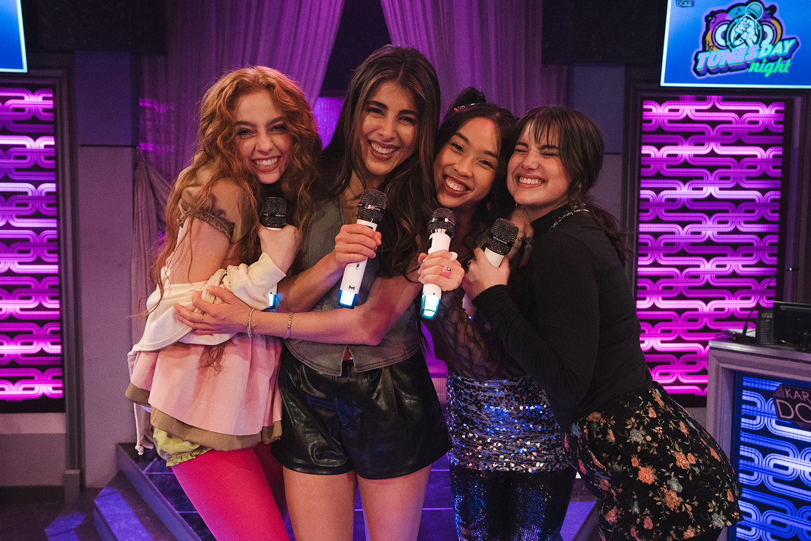 &Quot;Four Young Women Smiling And Hugging While Holding Microphones, Standing Together On A Colorful Karaoke Stage Decorated With Purple Lights And A &Quot;Tunes-Day Night&Quot; Sign, Suggesting A Lively And Fun Indoor Environment.&Quot;