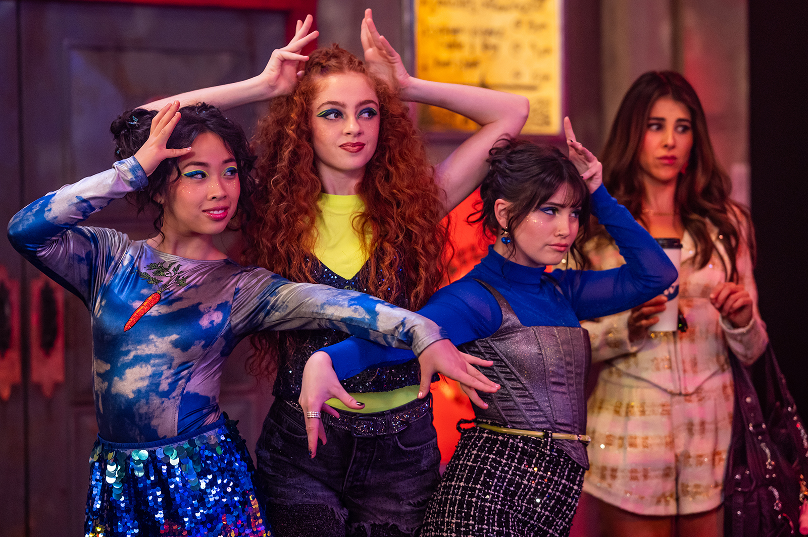 &Quot;Three Young Women In Colorful, Stylish Outfits Strike Dramatic Poses In A Lively Indoor Setting, While A Fourth Woman In A White Outfit Stands Behind Them, Watching With A Neutral Expression. The Environment Is Vibrant And Energetic.&Quot;