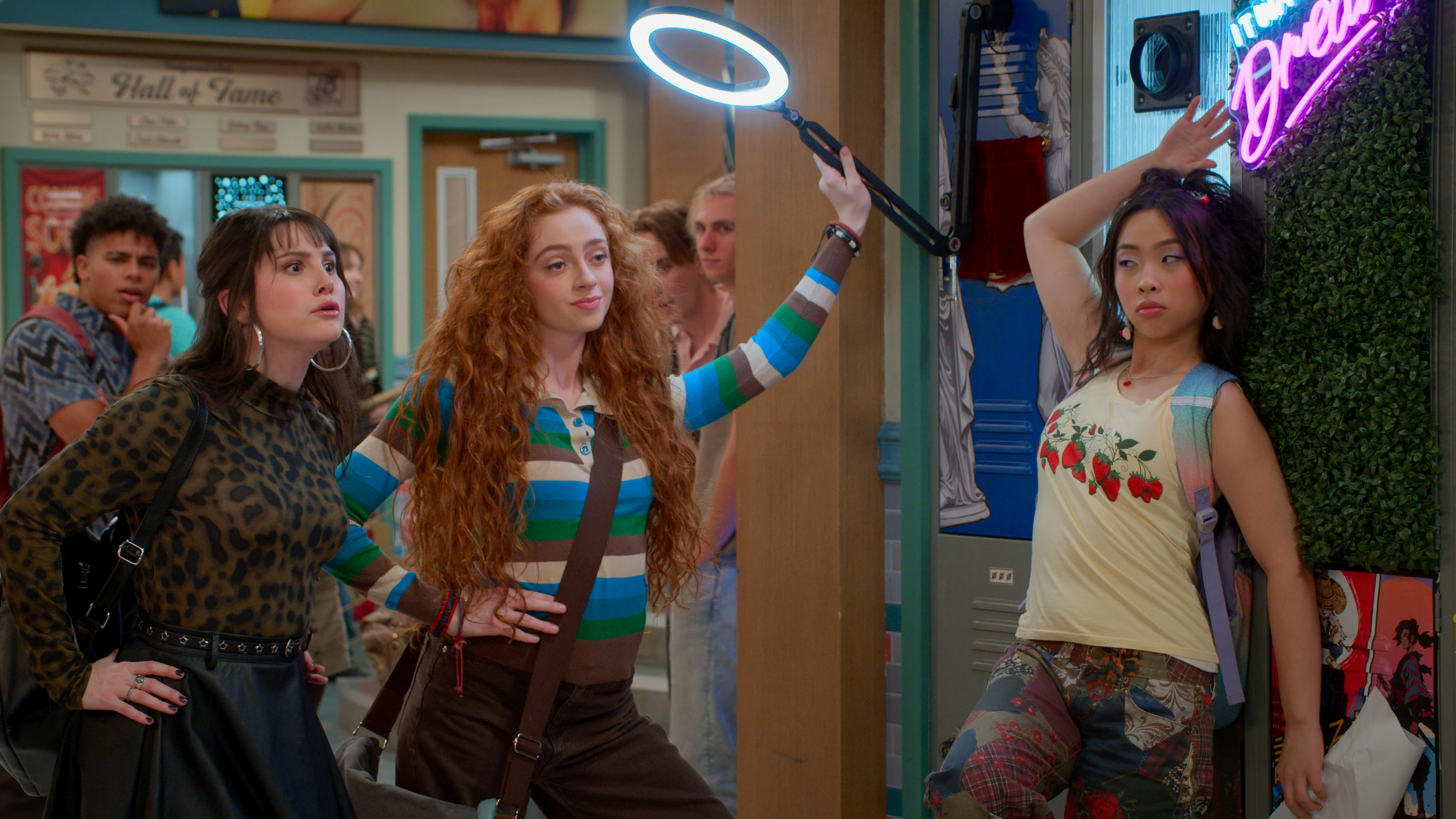 &Quot;Three Stylish Teens Pose Confidently In A Colorful School Hallway, One Holding A Ring Light, With Lockers, Posters, And A Neon Sign In The Background. Other Students Are Visible Further Down The Hall.&Quot;
