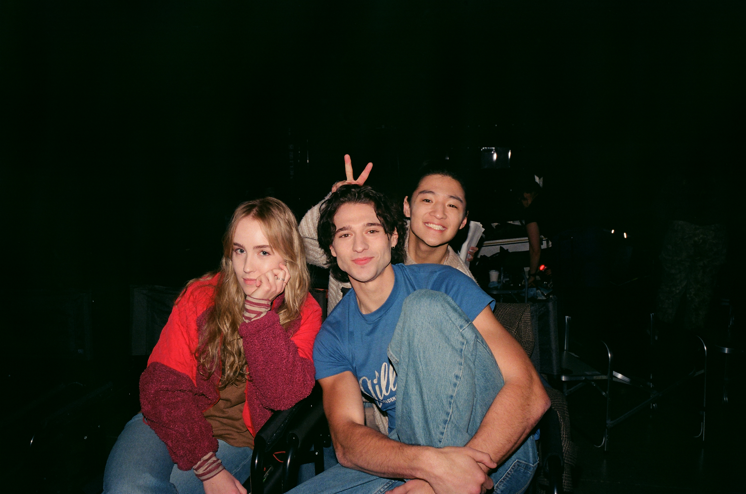 &Quot;Three Young Adults Sit Close Together In A Dimly Lit Room, Posing Casually. The Woman On The Left Has Long Blonde Hair And A Red Jacket, While The Two Men On The Right Smile, With One Making Bunny Ears Behind The Other.&Quot;