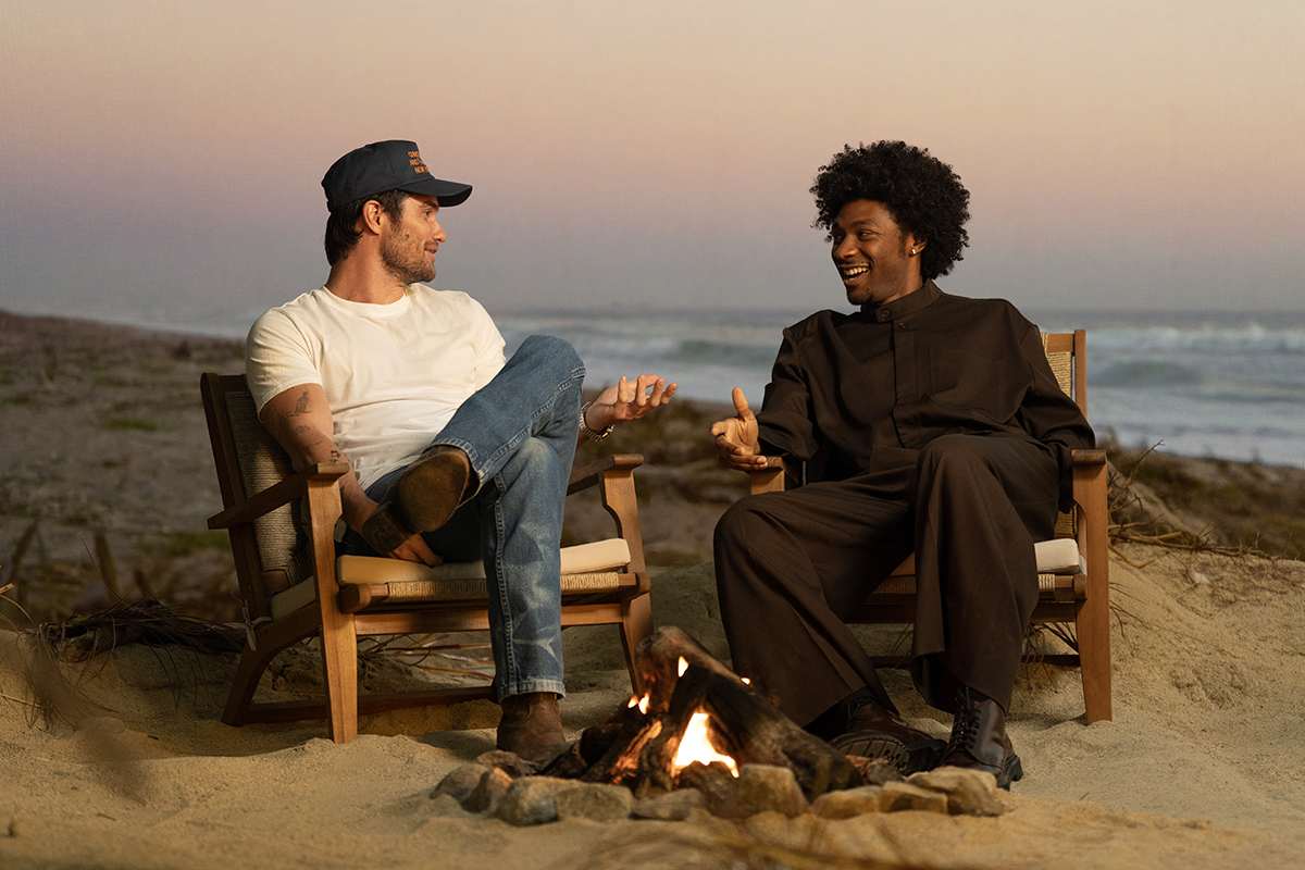 &Quot;Two People Sitting In Chairs On A Sandy Beach By A Campfire, Talking And Gesturing As The Sun Sets Over The Ocean In The Background, Creating A Relaxed And Friendly Outdoor Atmosphere.&Quot;