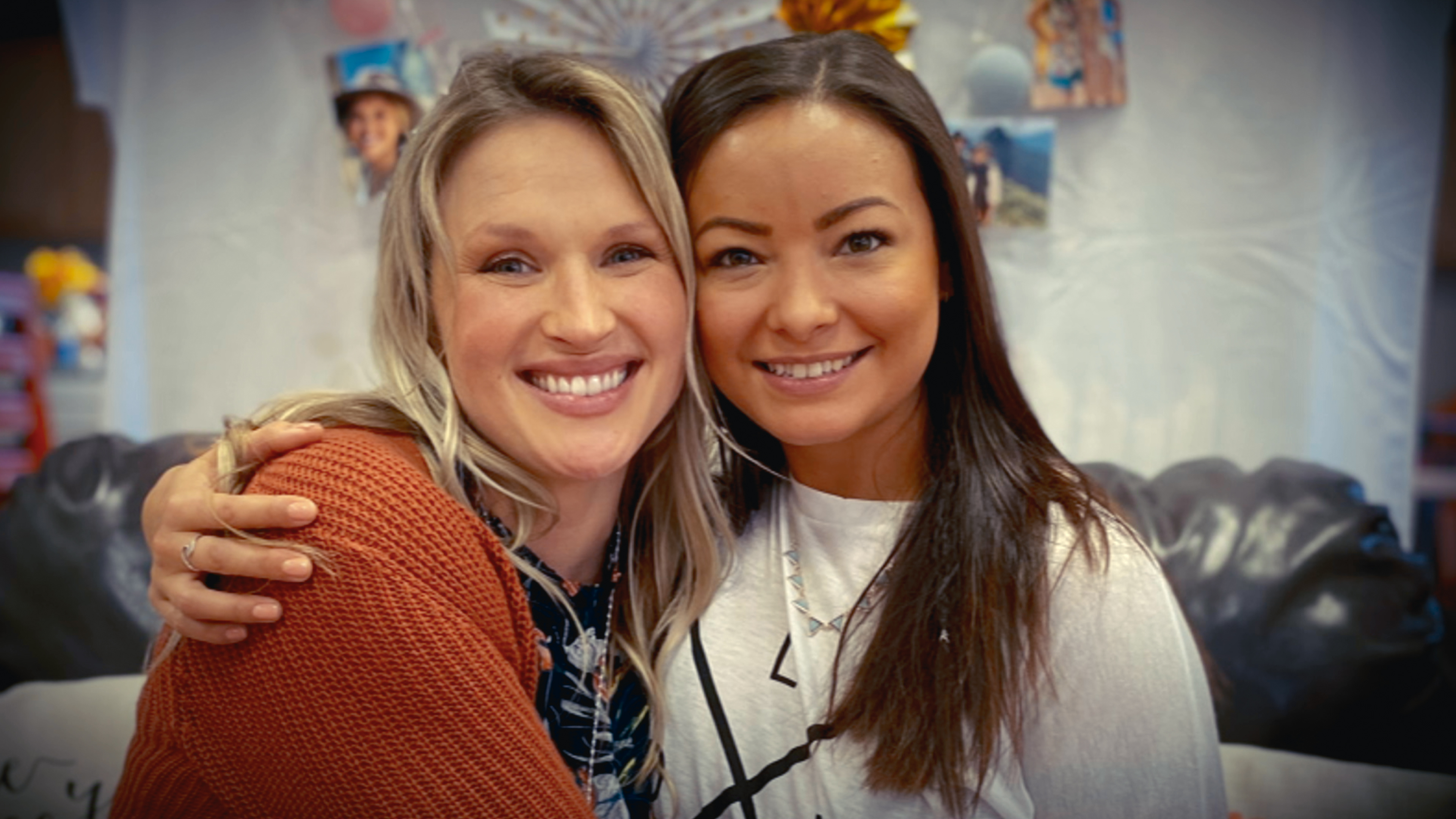 &Quot;Two Women Smiling And Sitting Close Together On A Black Couch, Embracing Each Other In A Cozy Indoor Setting With Photos And Decorations In The Background.&Quot;