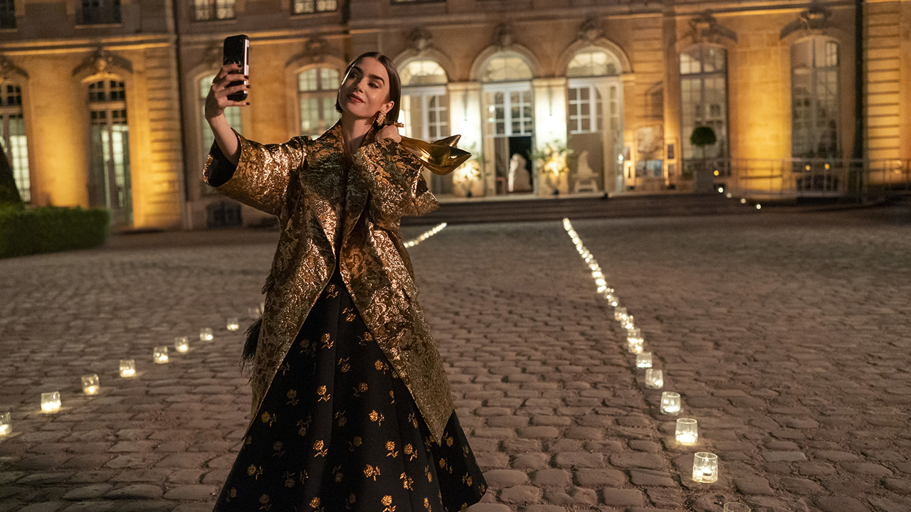 &Quot;Woman In A Gold Coat And Black Dress Takes A Selfie With Her Phone At Night In A Candlelit Courtyard Outside An Elegant, Well-Lit Building With Large Windows And Cobblestone Ground.&Quot;