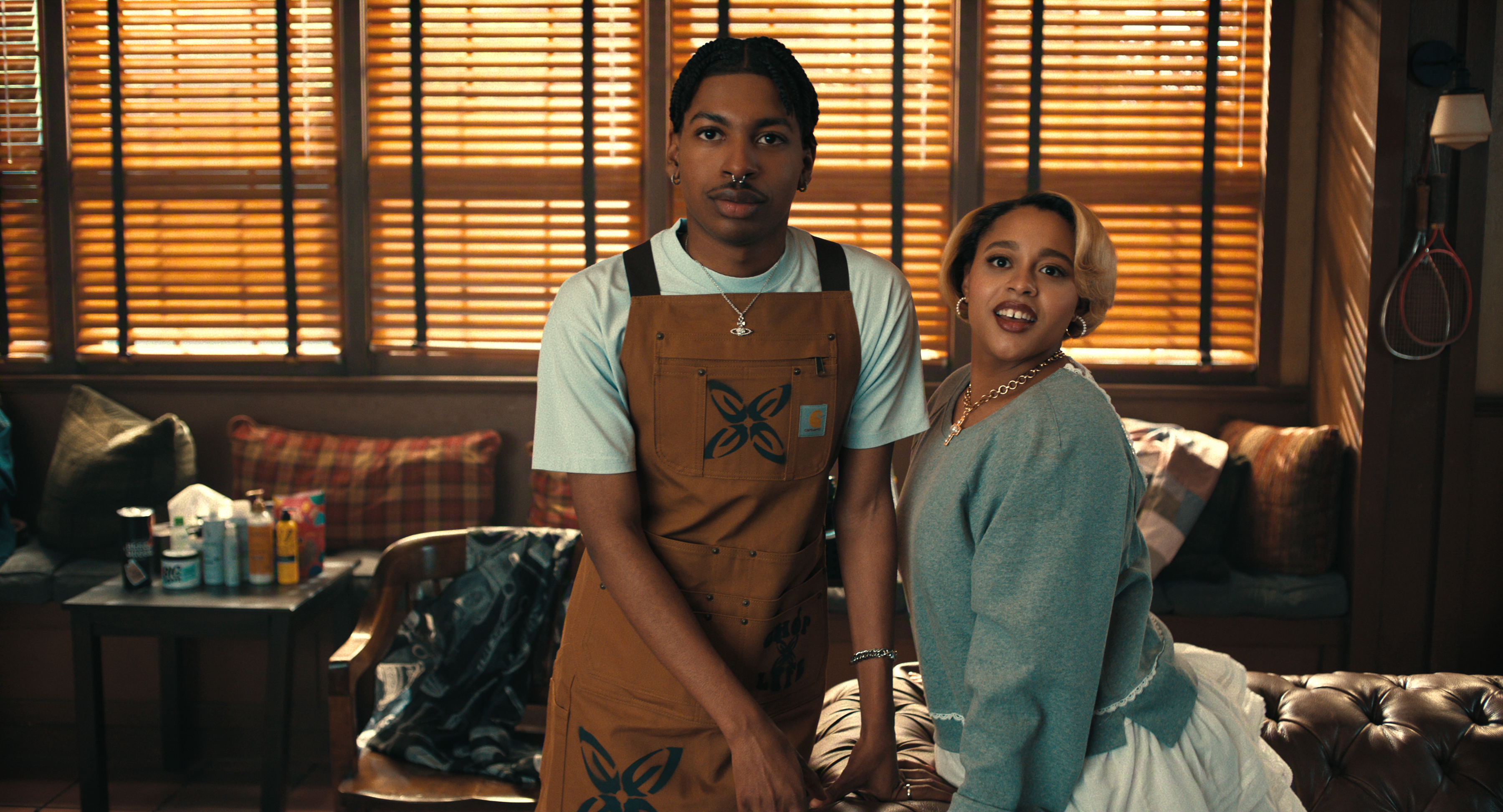 &Quot;Two People Pose Together In A Cozy, Sunlit Room With Wooden Blinds, Patterned Pillows, And Beauty Products On A Nearby Table. One Person Wears An Apron, Suggesting A Salon Or Styling Environment.&Quot;