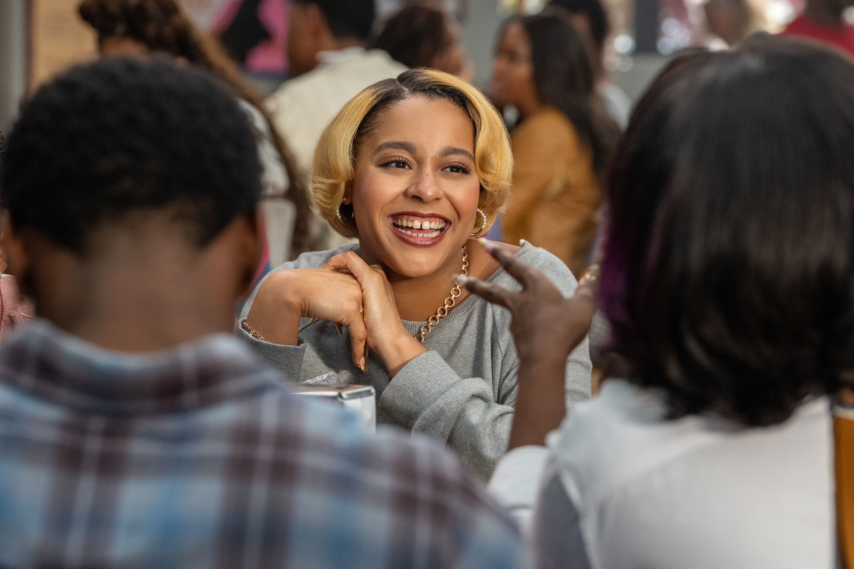 &Quot;Smiling Woman With Short Blonde Hair Sits At A Table, Talking And Laughing With Friends In A Lively, Casual Restaurant Or Café Setting With Other Diners In The Background.&Quot;