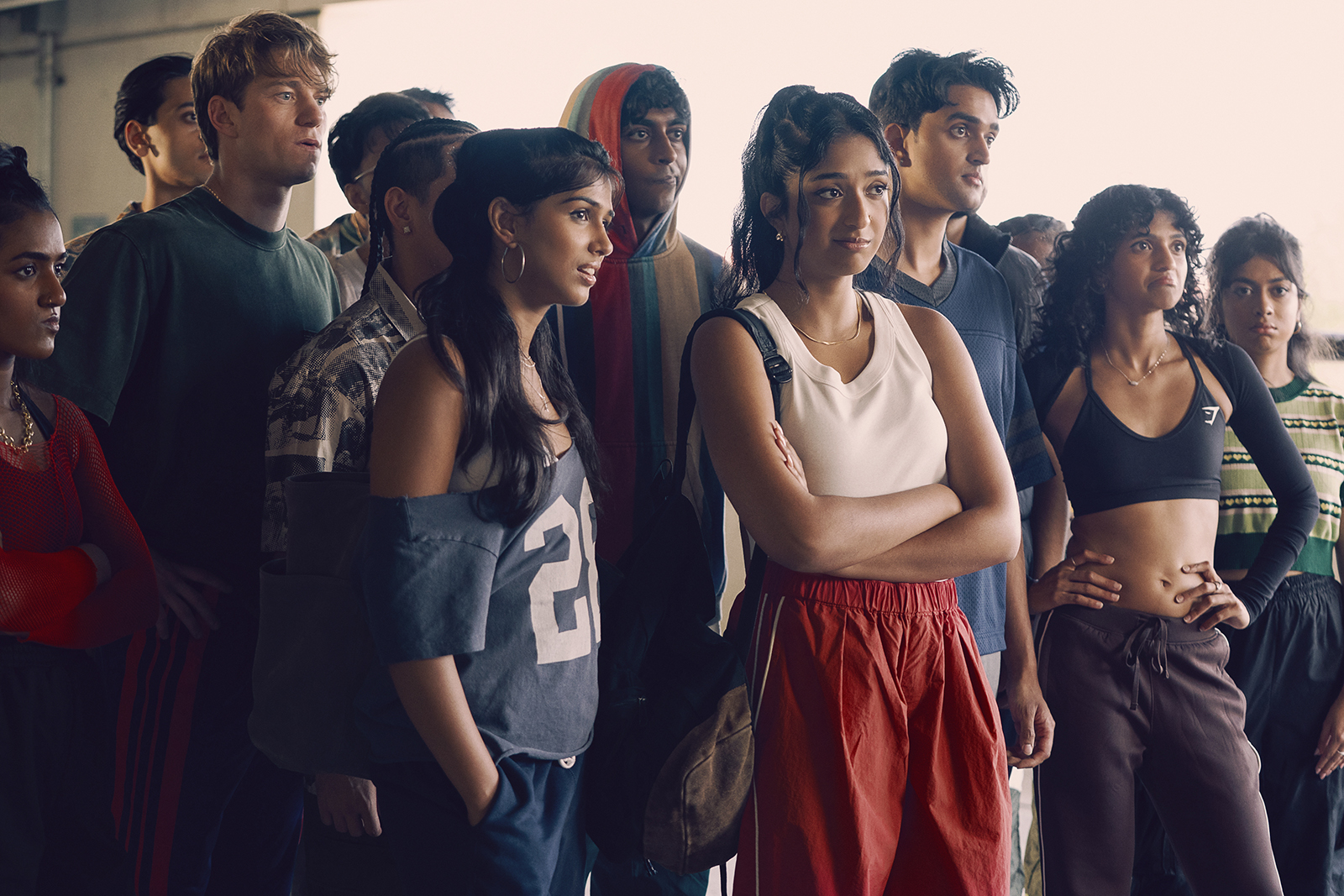 "A diverse group of young adults in sporty, casual clothing stand together indoors, appearing focused and attentive, possibly listening to someone or watching an event in a well-lit urban setting."