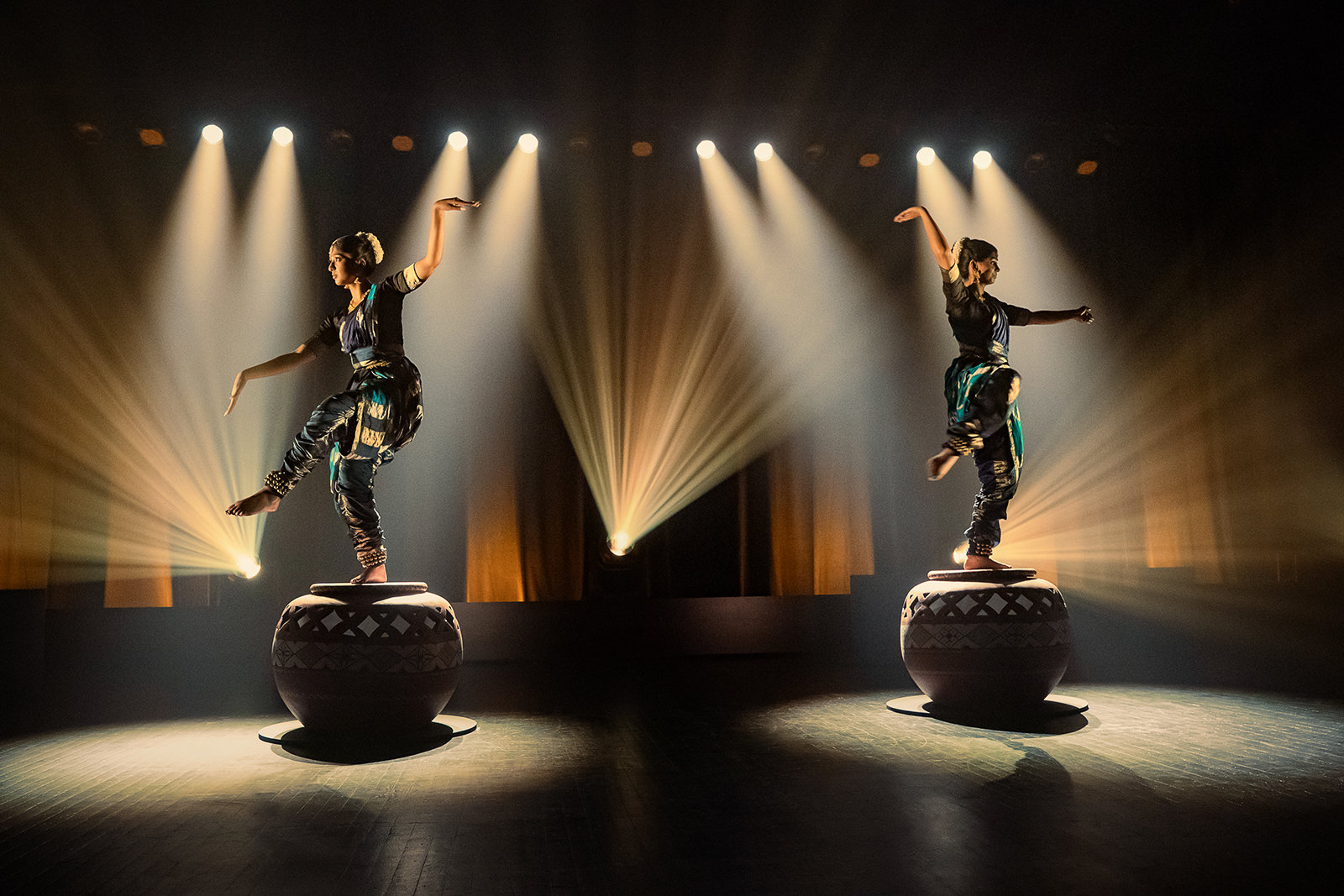 "Two performers in traditional attire balancing on large decorated drums on a stage, dramatic spotlights and beams illuminating them in a dark theater setting."