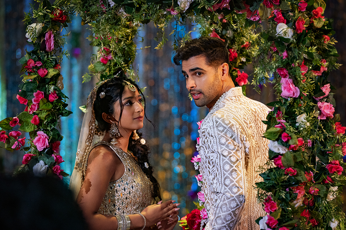 &Quot;A Couple In Traditional Indian Attire Stands Under A Vibrant Floral Arch At A Decorated Event, Possibly A Wedding Or Celebration, Surrounded By Colorful Flowers And Shimmering Blue Background Lights.&Quot;