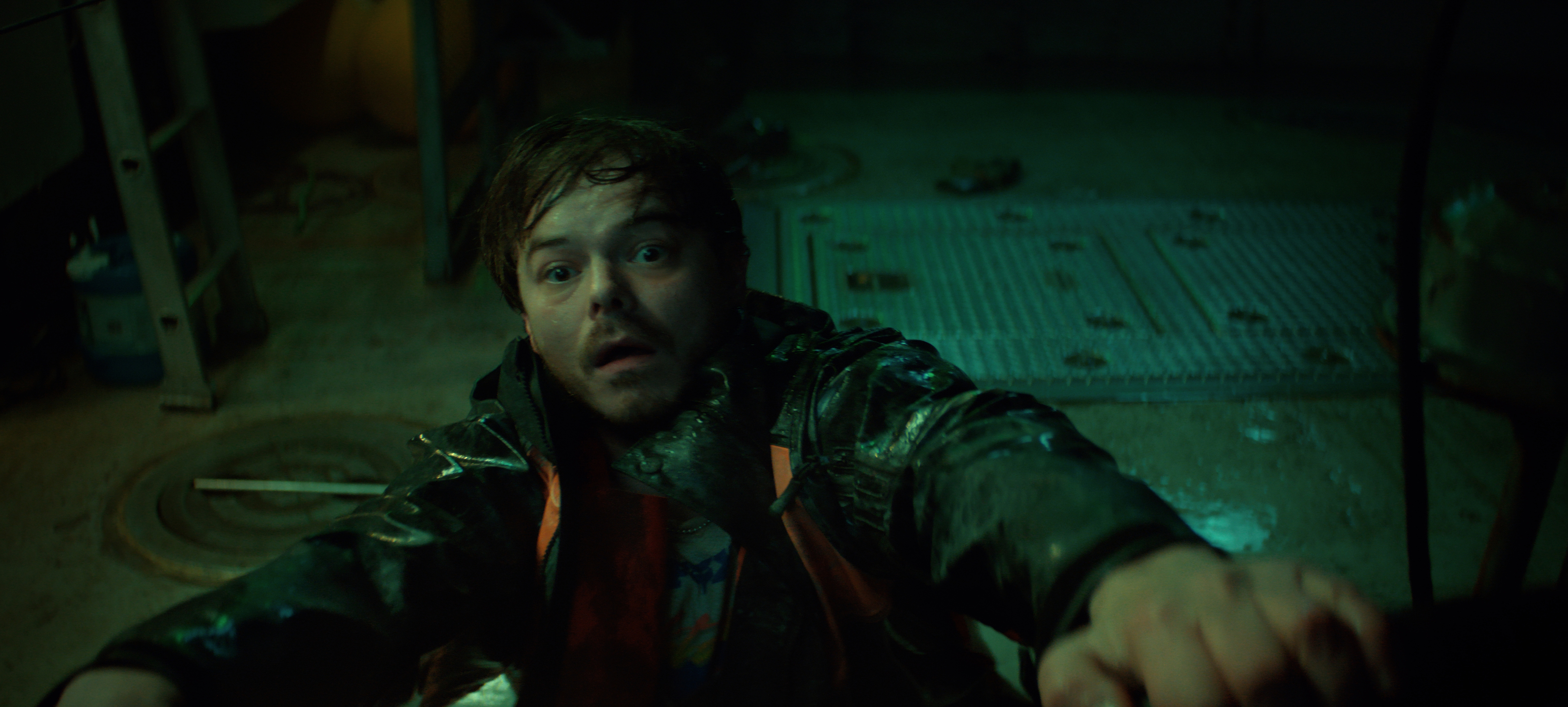 "A frightened man in a raincoat clings to a metal railing inside a dimly lit, industrial room with wet floors and scattered tools, suggesting tension or danger in the environment."
