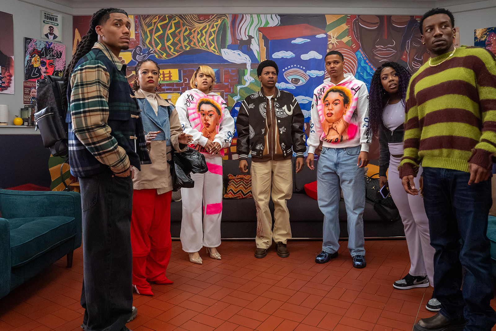 "Seven young people stand in a colorful room with bold artwork, looking surprised or worried. The backdrop features graffiti-style murals and a sofa, suggesting a casual, creative environment."