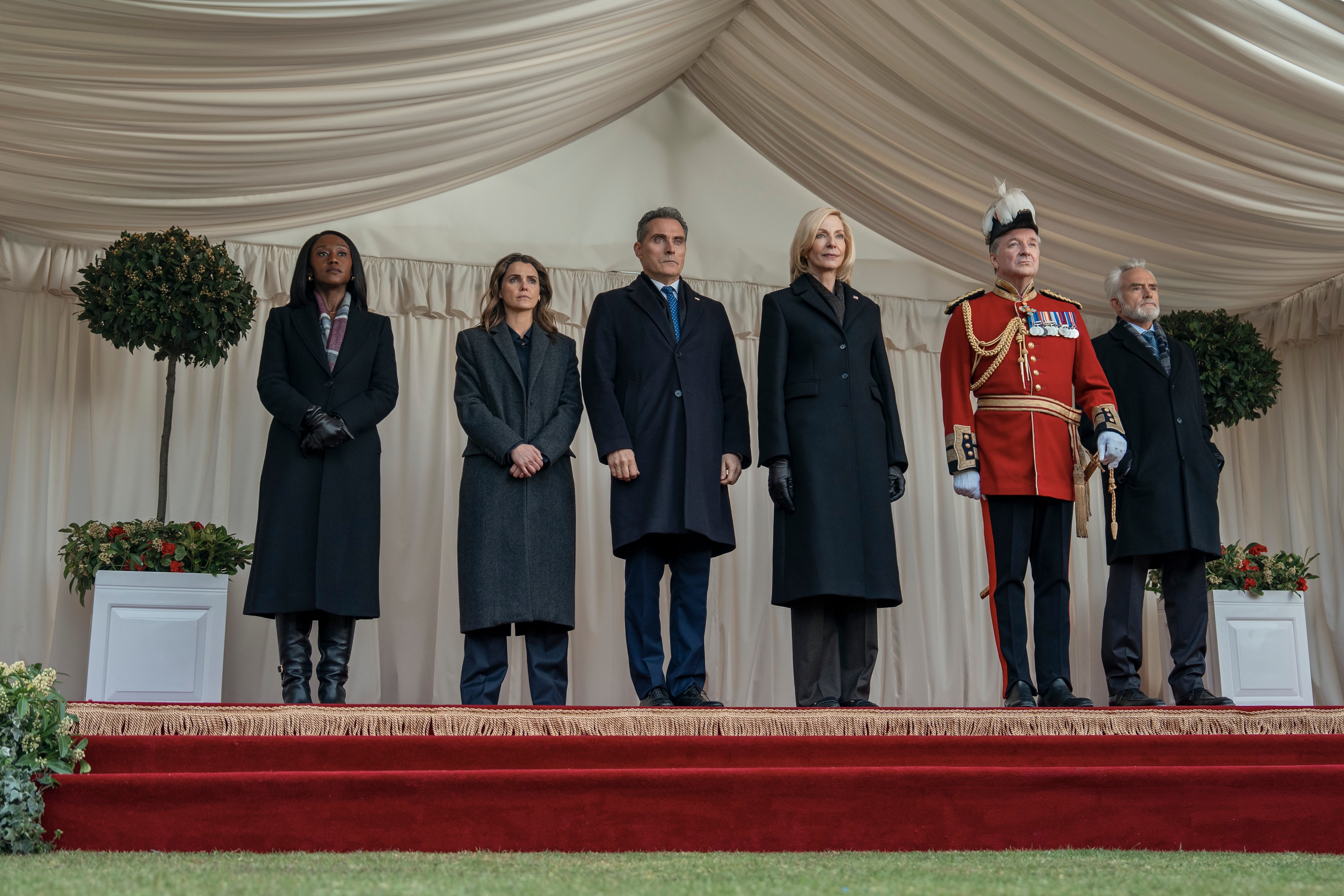 "Six formally dressed figures stand on a red-carpeted stage under a white draped umbrella, surrounded by potted plants; One wears a ceremonial military uniform, the others wear dark coats, and appear to be attending a formal or formal outdoor event."