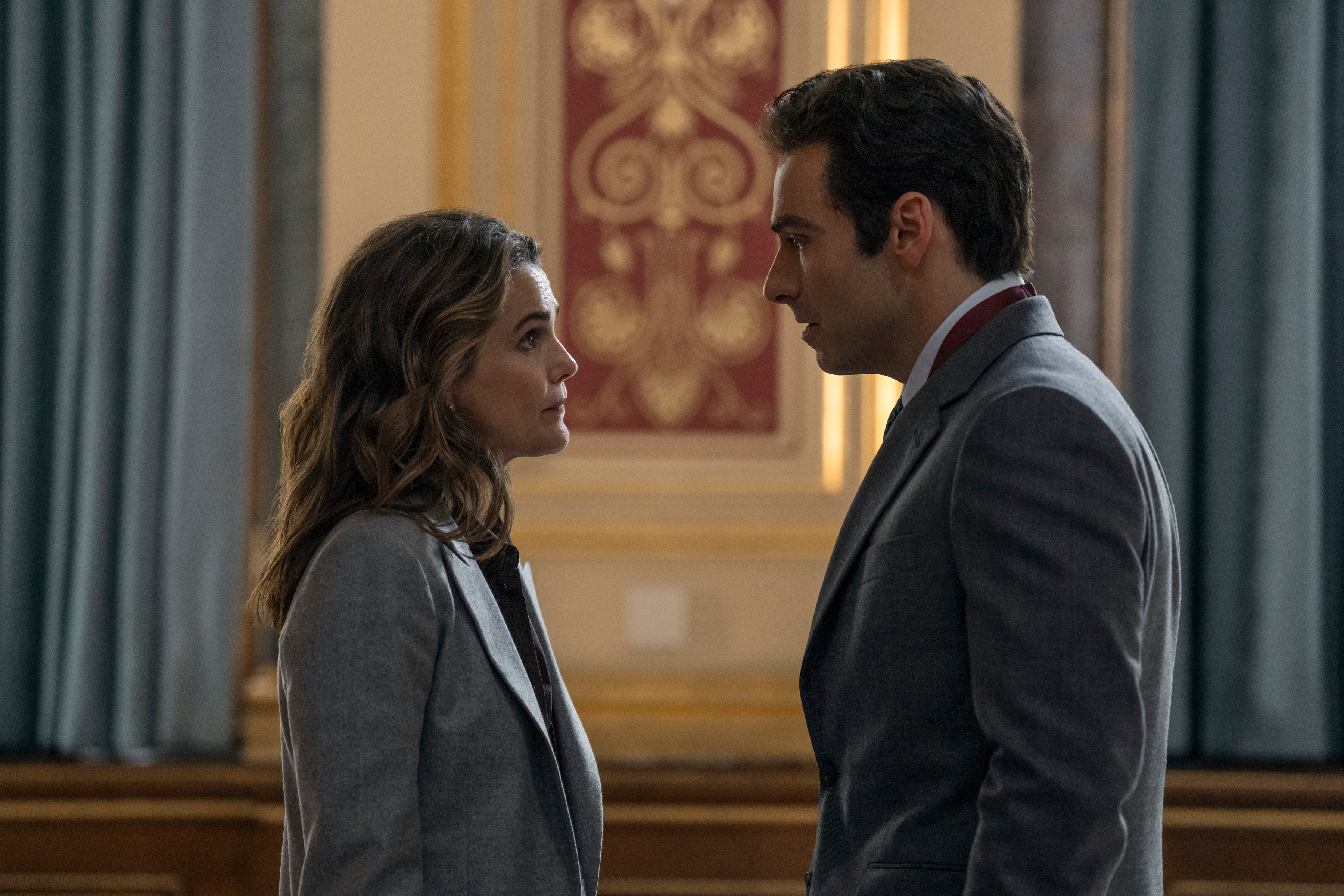 "Two people in business attire stand facing each other in an ornate formal room with blue curtains and patterned wall panels, appearing to be engaged in a serious conversation."