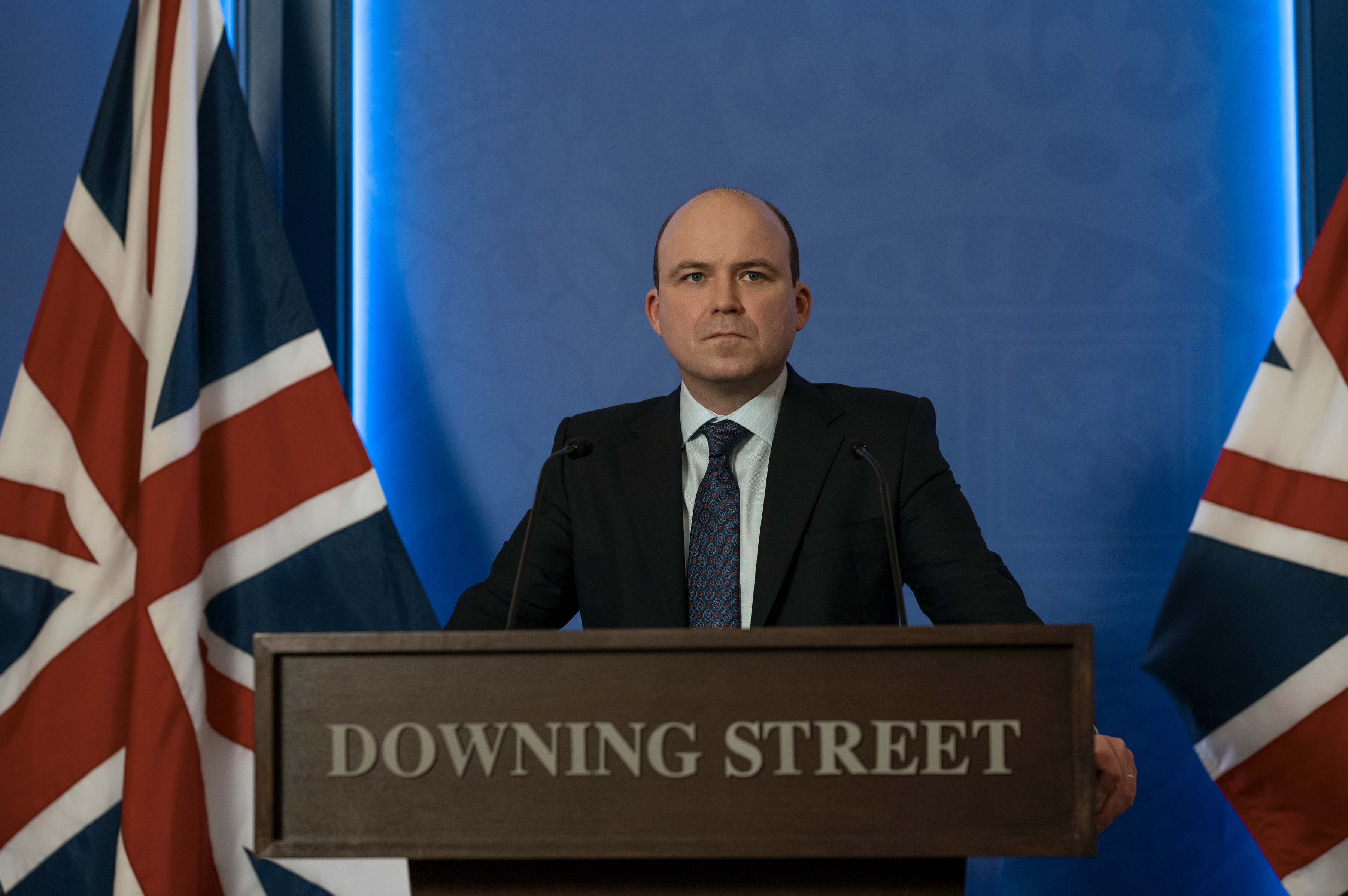 "A man in a suit stands on the Downing Street stage, flanked by two British flags, making a statement at an official press conference with a blue background".