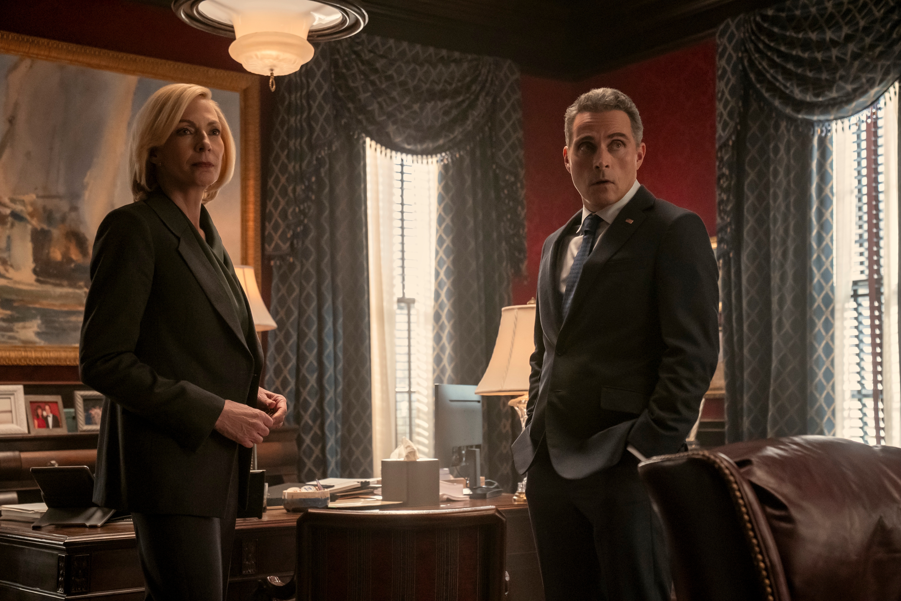 "Two people in formal attire stand in an elegant, dimly lit office with large windows, curtains, and classic decor. They seem to be in a serious conversation or meeting."