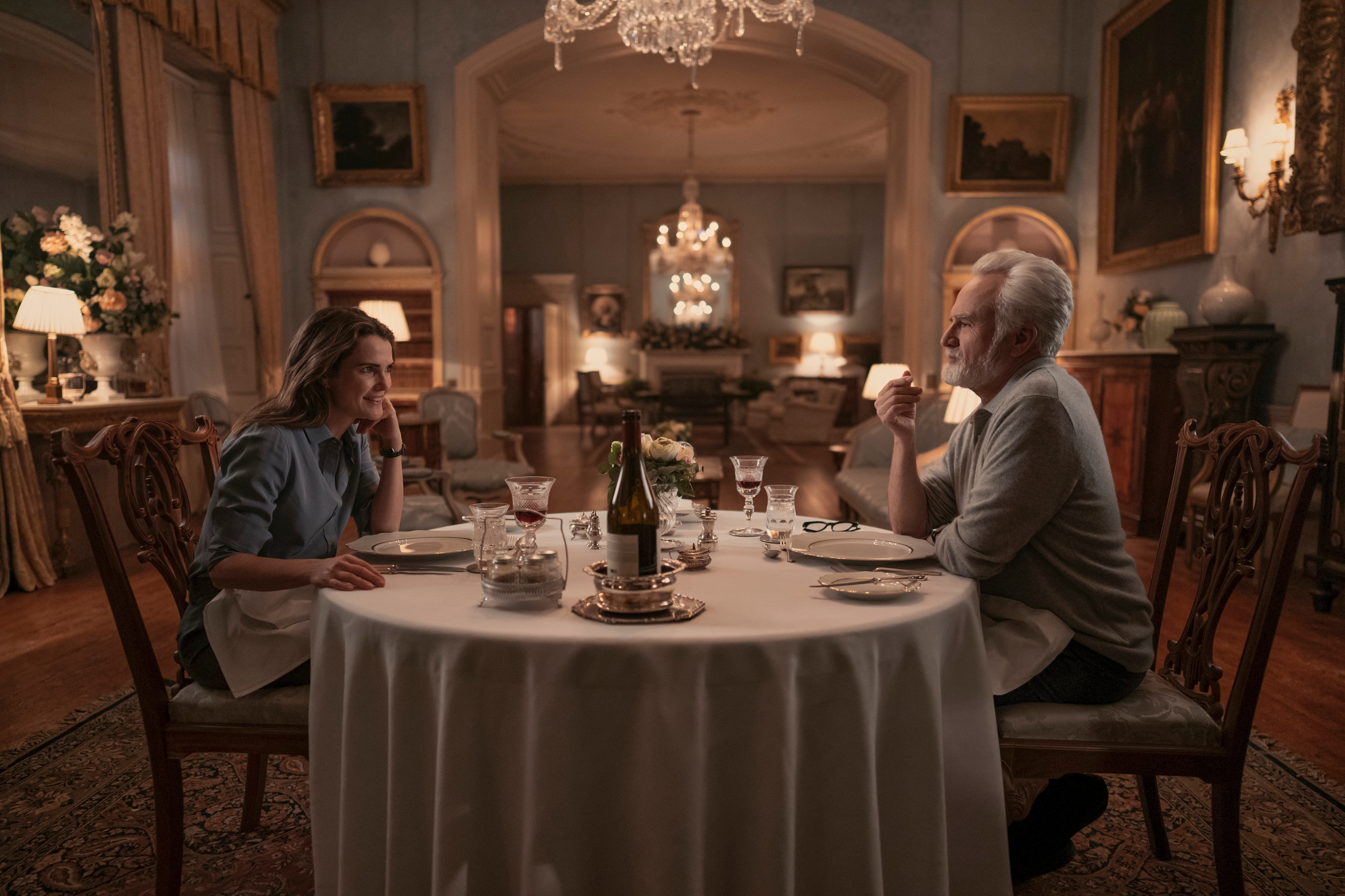 "Two people sit opposite each other at an elegantly set round dining table in a luxurious, warmly lit room with chandeliers, framed paintings and ornate decor, suggesting an intimate or formal dinner setting."