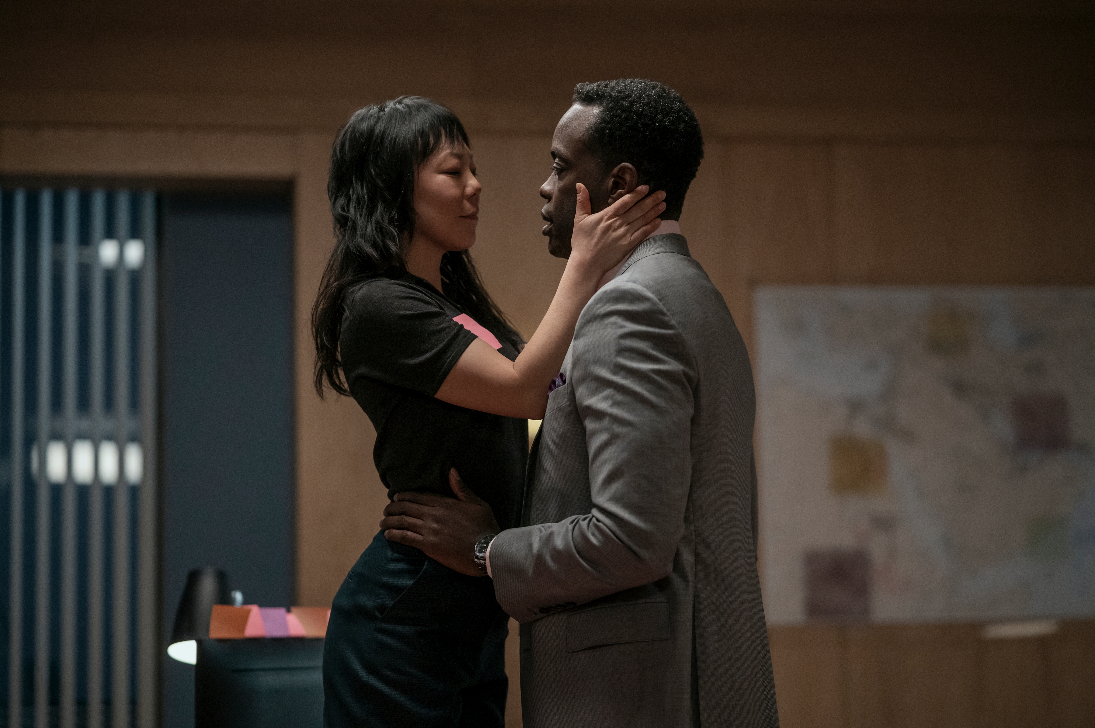 "A woman and a man embrace closely in an office, surrounded by wooden walls, a desk with papers, and a map on the wall, conveying an intimate moment between colleagues or partners."