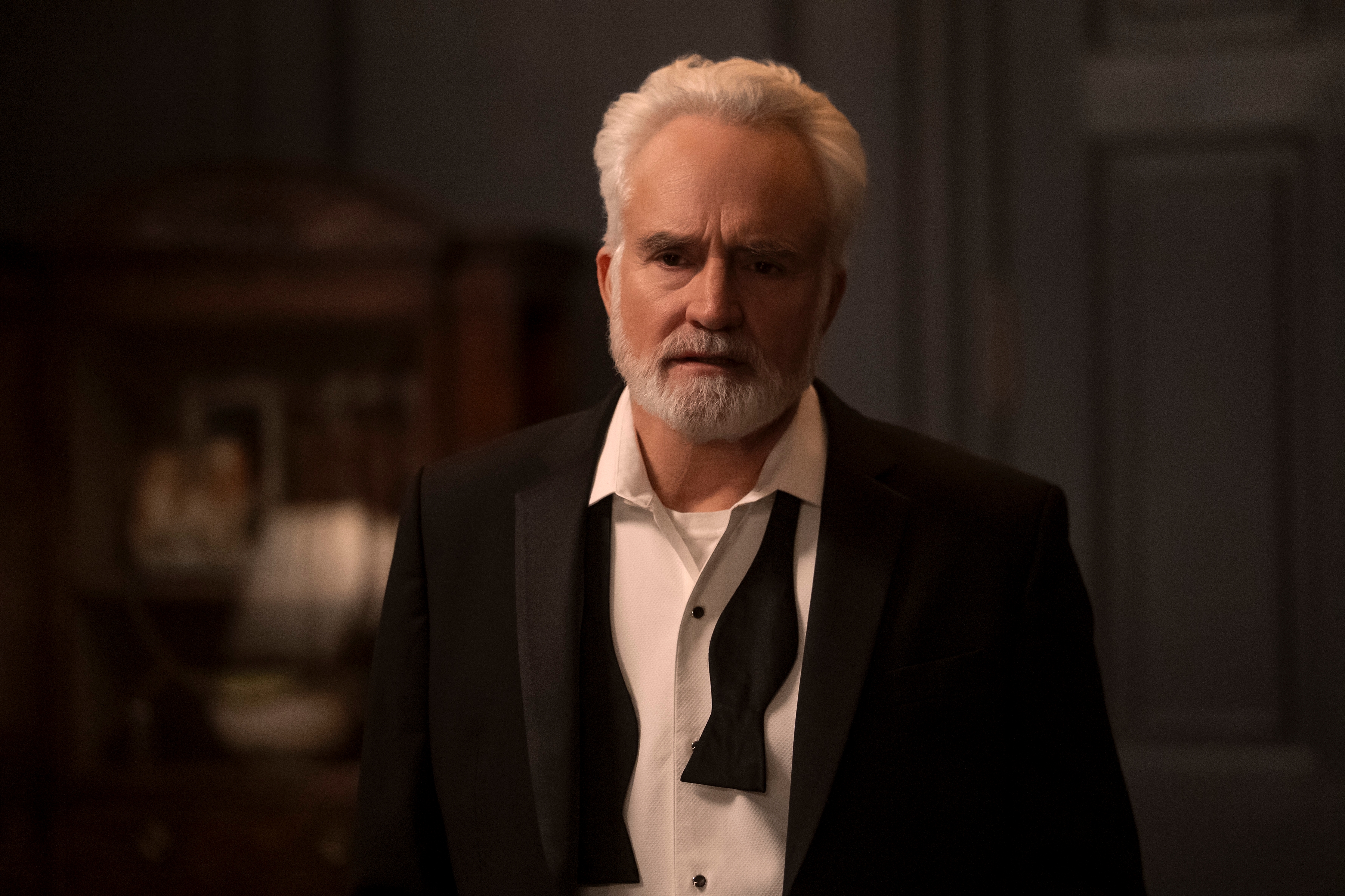 "An older man with white hair and a beard wearing a business suit with an untied tie, standing indoors in a dimly lit formal room with dark walls and a closet in the background."