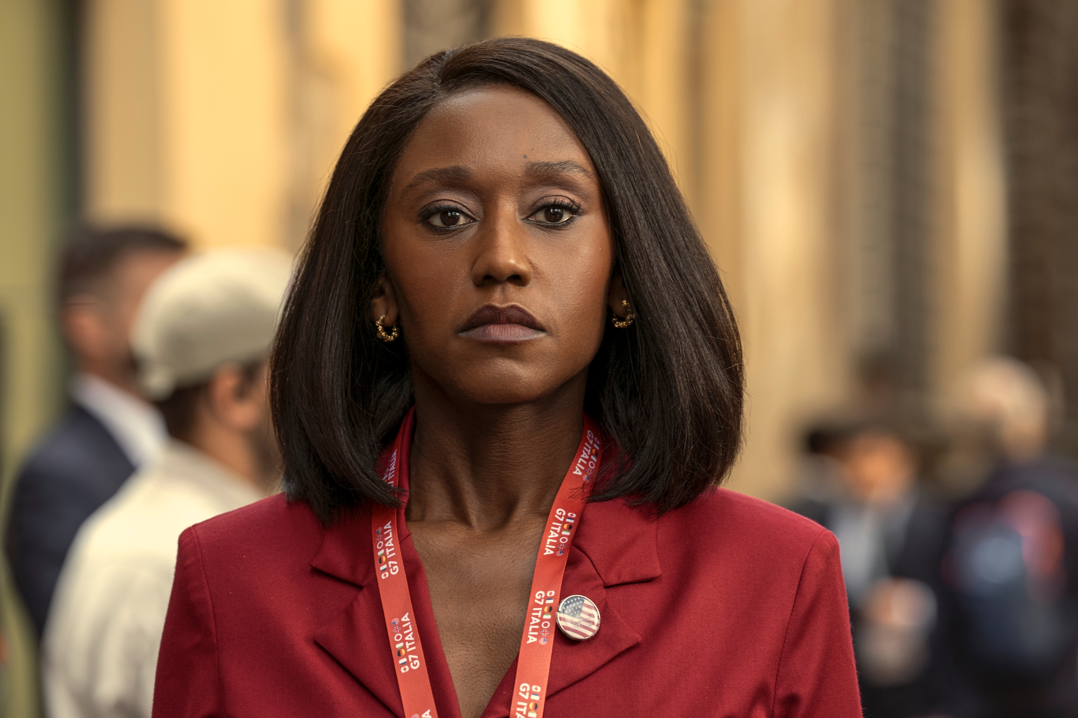 "A woman wearing a red jacket and a press badge stands outside in a busy urban environment, looking serious. People and buildings are blurred in the background, suggesting a public event or news coverage."