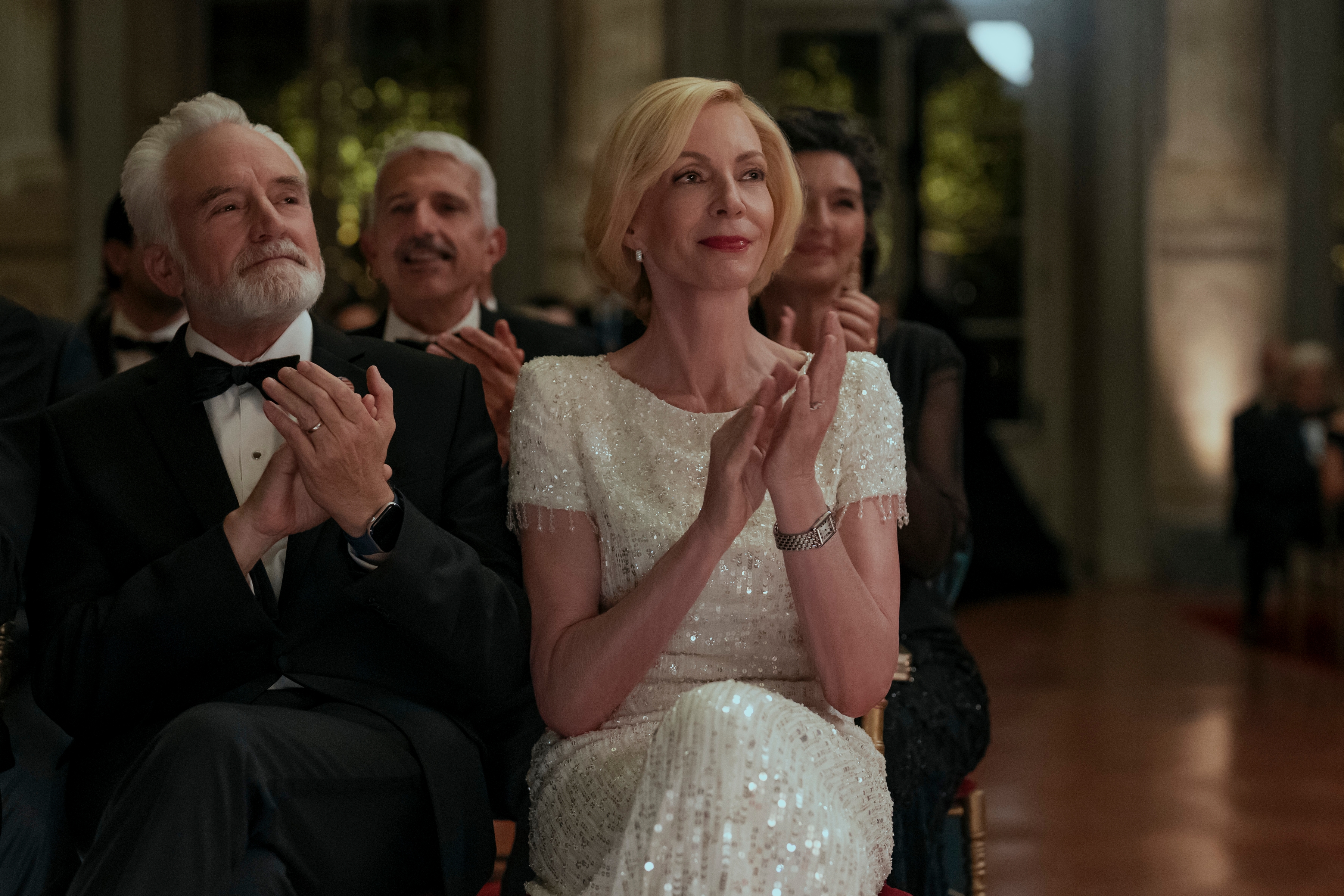 "Elegant people in formal attire sit and clap at an indoor event, perhaps a party or celebration, with warm lighting and ornate decor in the background."