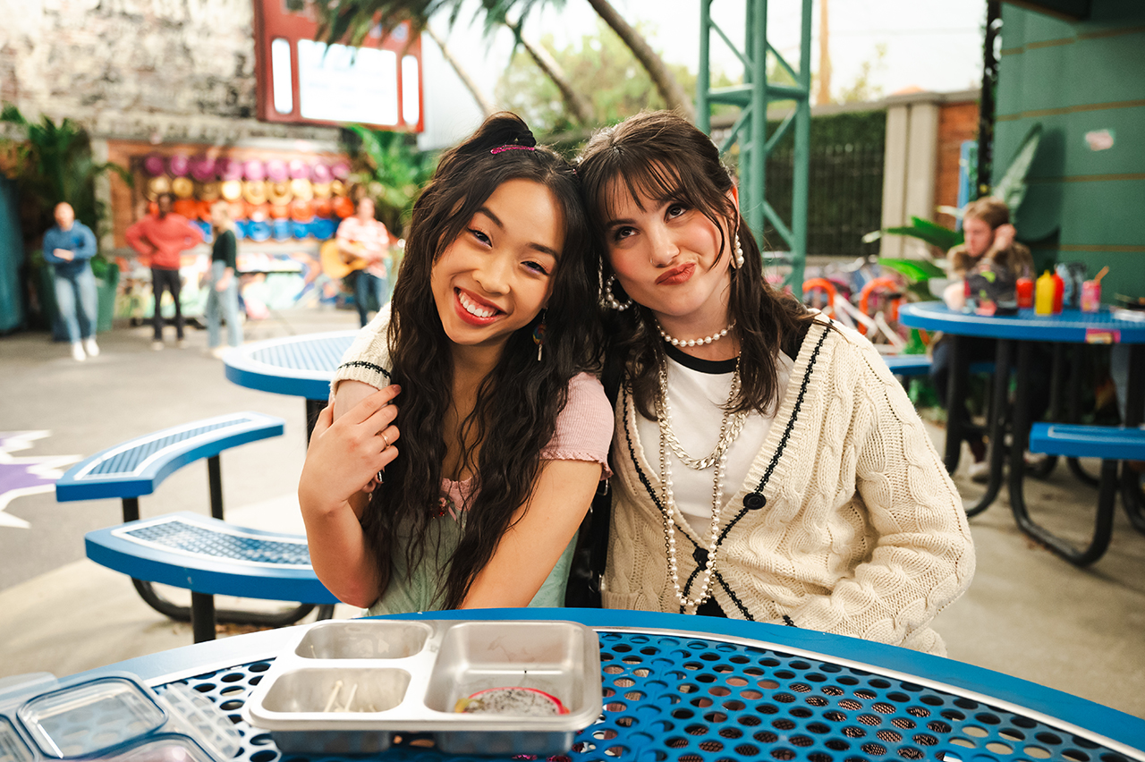 &Quot;Two Teenage Girls Sit Closely Together Smiling At A Blue Outdoor Picnic Table In A Lively, Colorful Amusement Park Or Playground Setting, With Other People And Games Visible In The Background.&Quot;