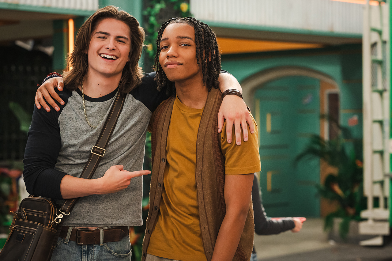 &Quot;Two Smiling Teenage Boys Stand Together Indoors In A Brightly Lit Room, One With His Arm Around The Other's Shoulder And The Other Pointing At His Friend. There Are Plants And A Green Door In The Background, Creating A Casual Atmosphere.&Quot;