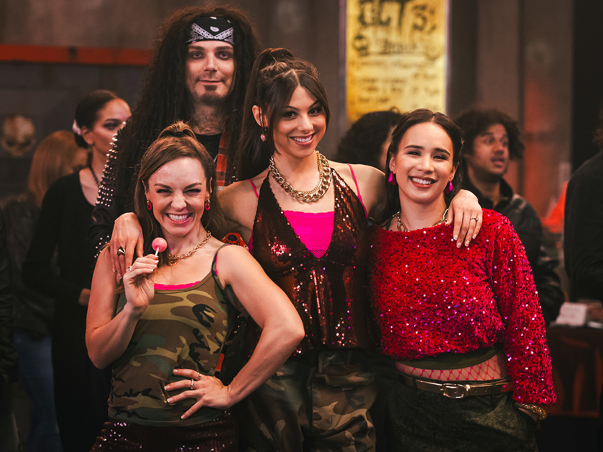 &Quot;Three Smiling Women, Wearing Colorful And Shiny Outfits, Pose Together In A Lively Indoor Setting With A Crowd And A Man In A Wig And Costume Behind Them, Suggesting A Party Or Festive Event.&Quot;
